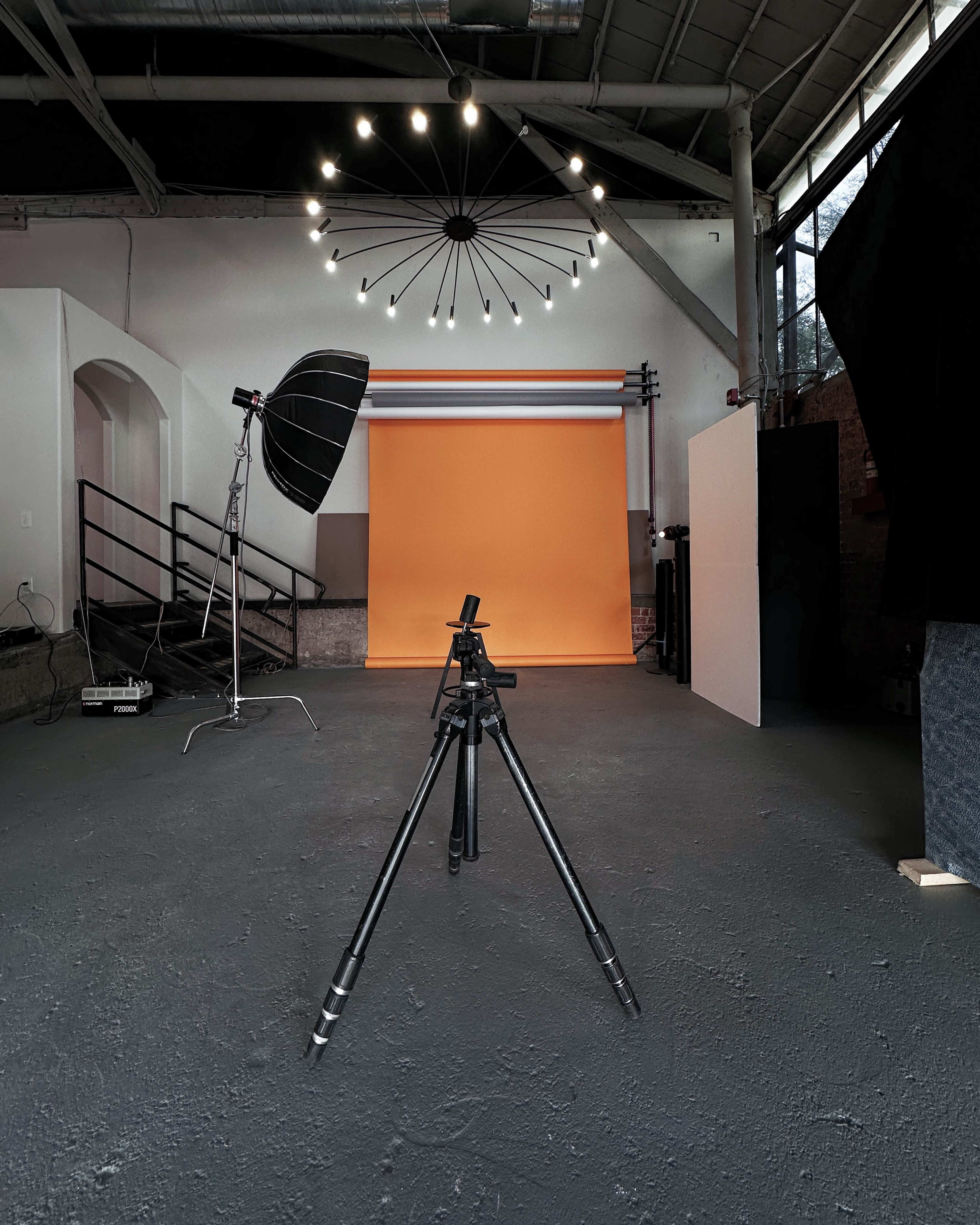 A photography studio with a black tripod positioned on a gray floor, facing an orange backdrop and illuminated by a circular light fixture.