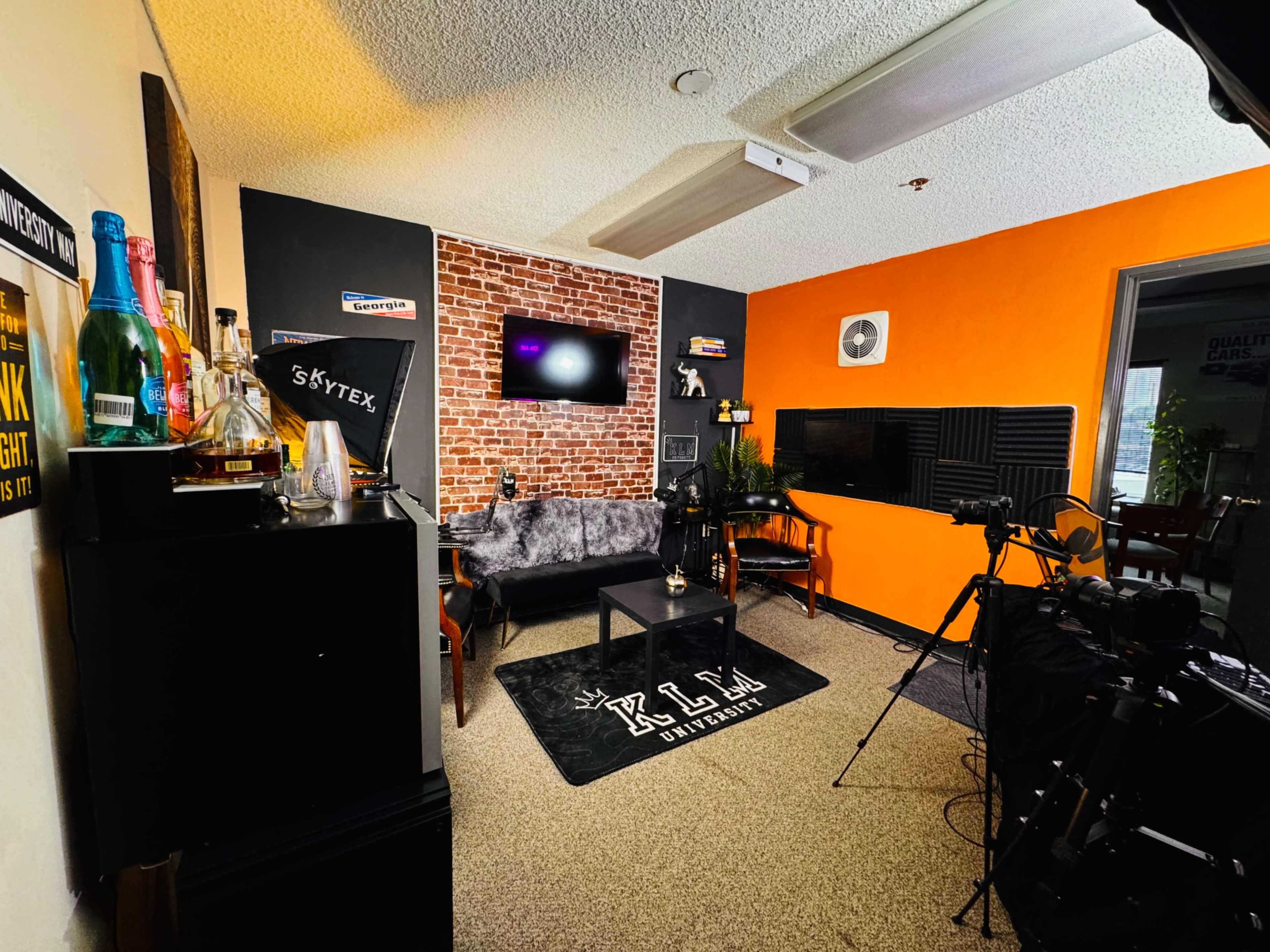 The image shows a cozy room with a brick accent wall, a black couch, and a television, featuring a setup for video recording with a camera and microphone on tripods.