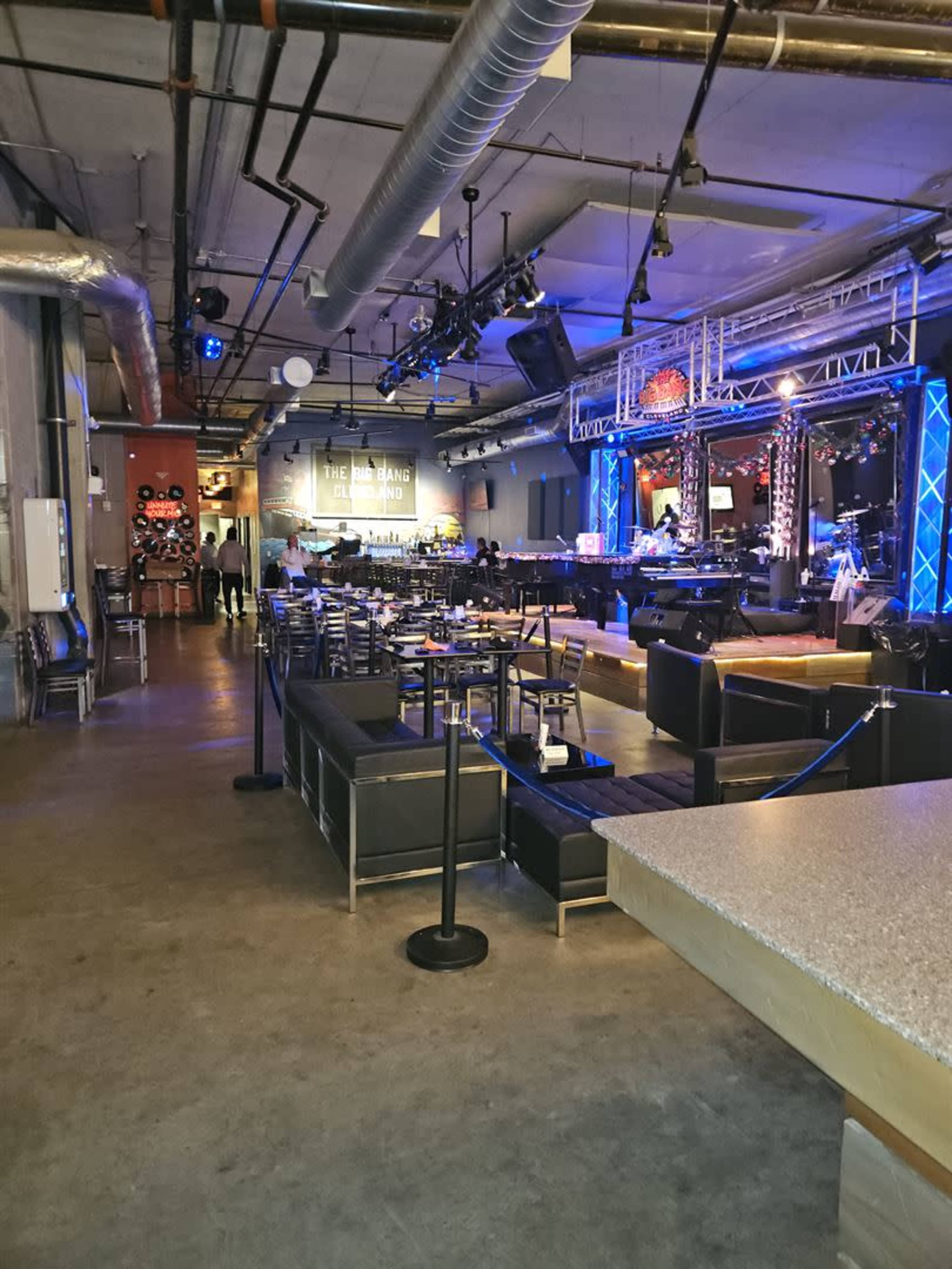 A spacious restaurant or bar interior with tables, chairs, and a stage area lit by colorful lights.