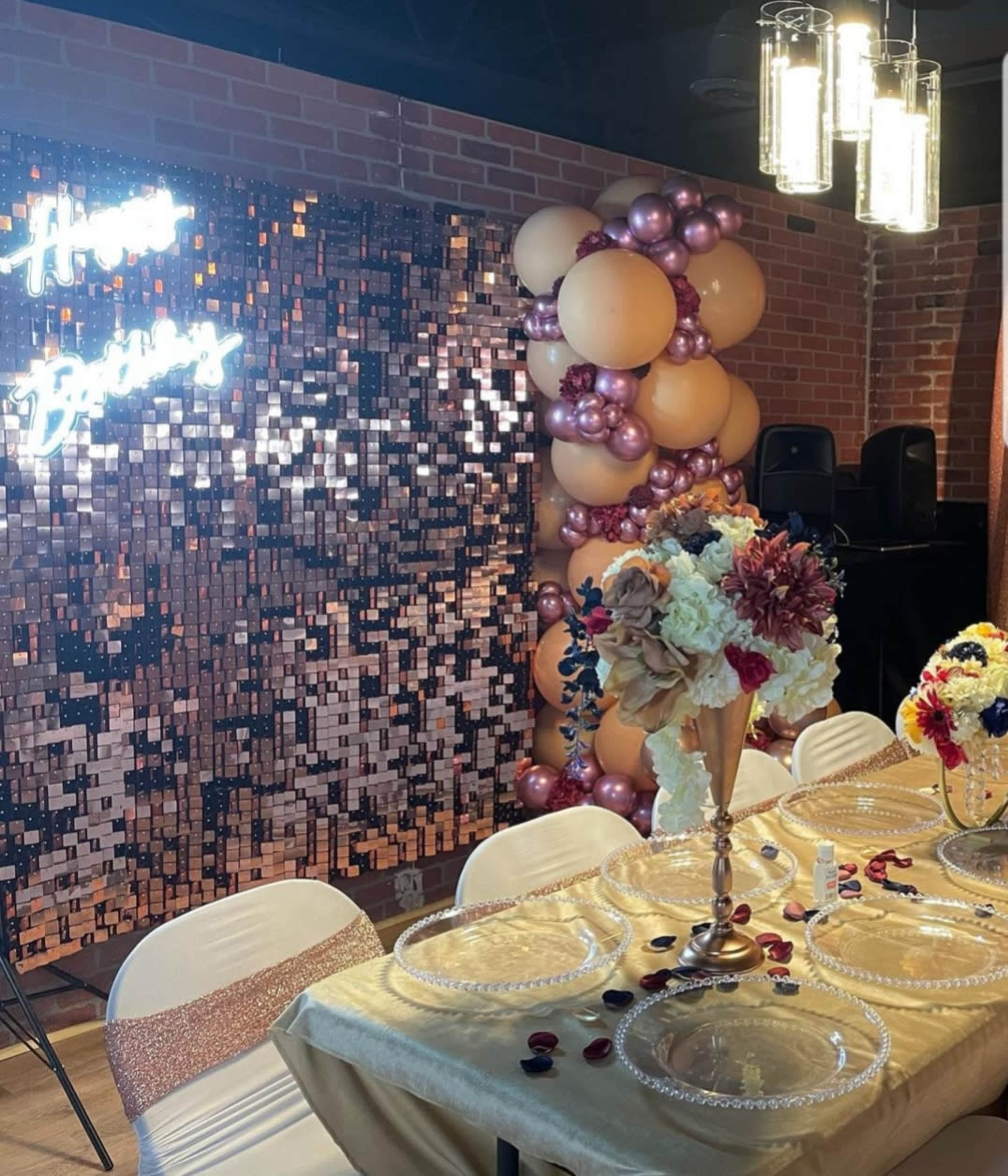 A birthday celebration setup featuring a decorated table with floral centerpieces, a shiny backdrop, and a balloon arch.