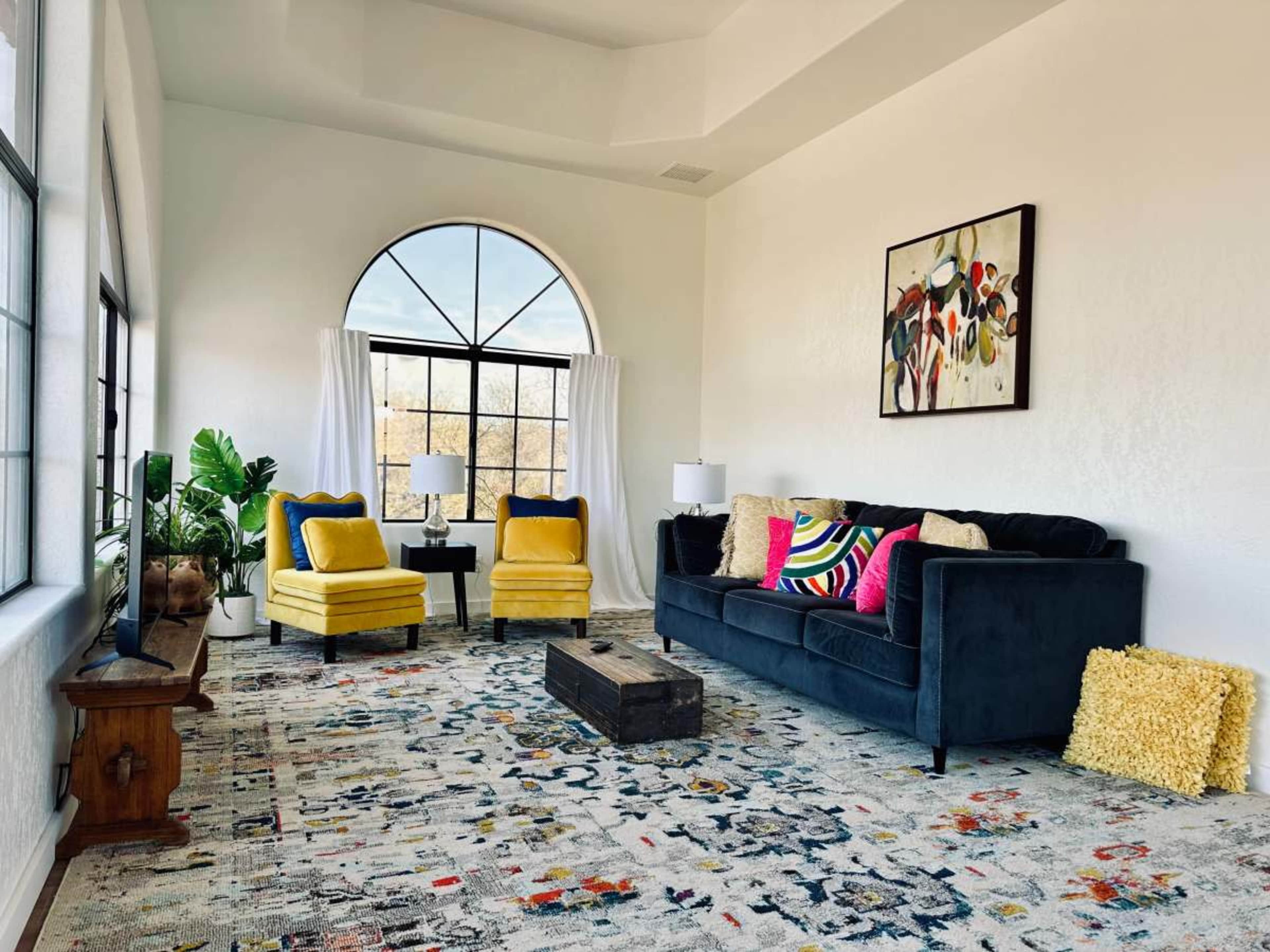 A bright living room features a dark sofa, two yellow armchairs, a wooden coffee table, and large windows with sheer curtains.