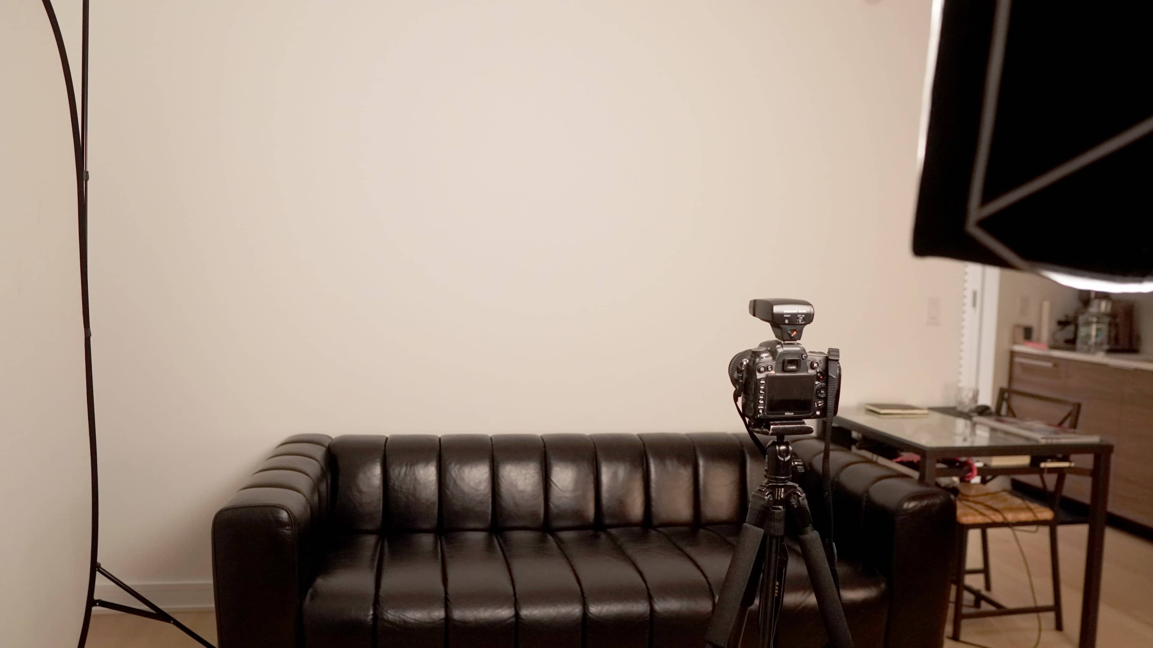 A black leather couch is positioned in front of a plain wall with a camera tripod set up nearby.