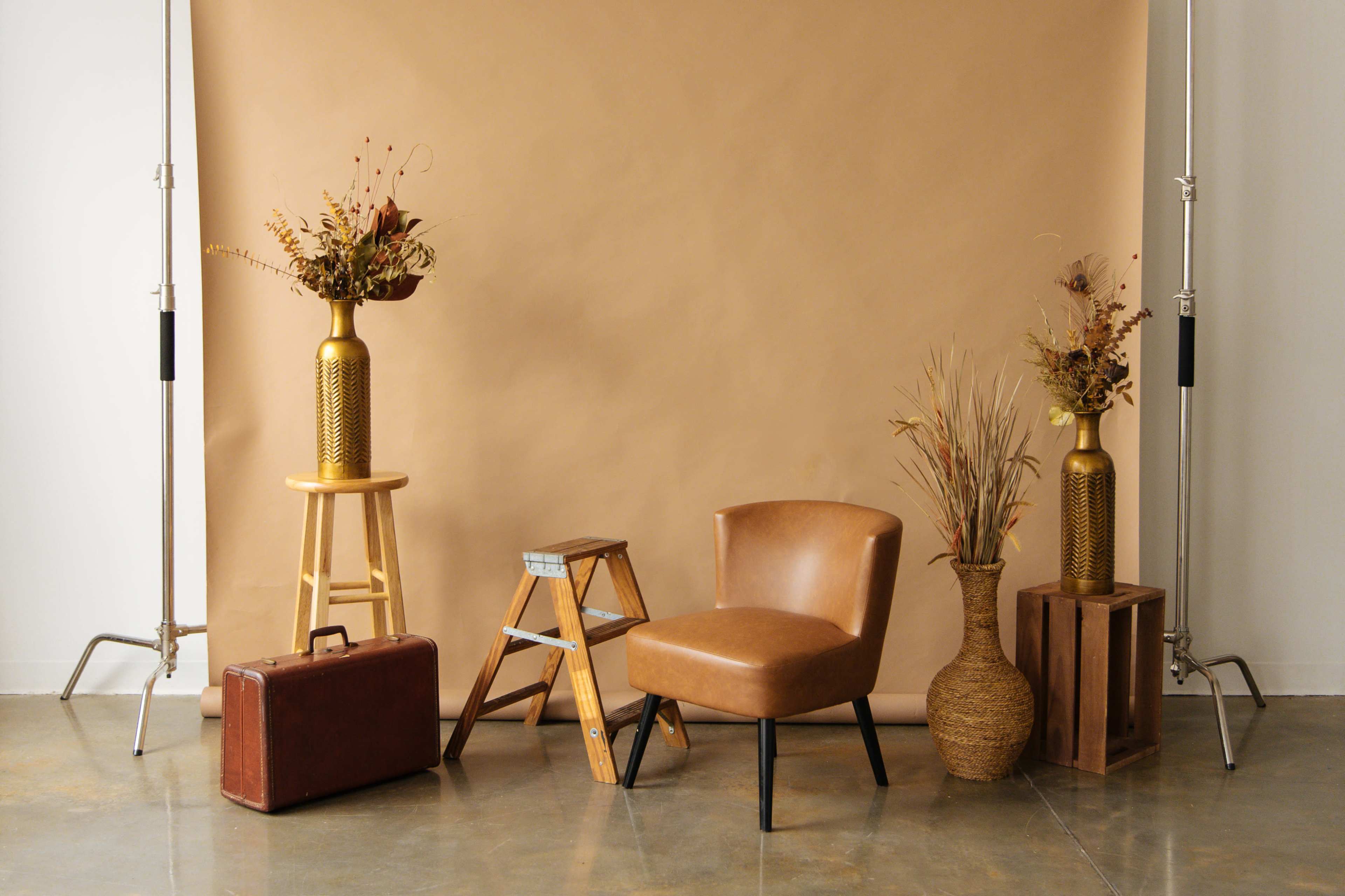 A styled scene with a brown leather chair, a wooden ladder, various vases with floral arrangements, a vintage suitcase, and a wooden crate against a beige backdrop.