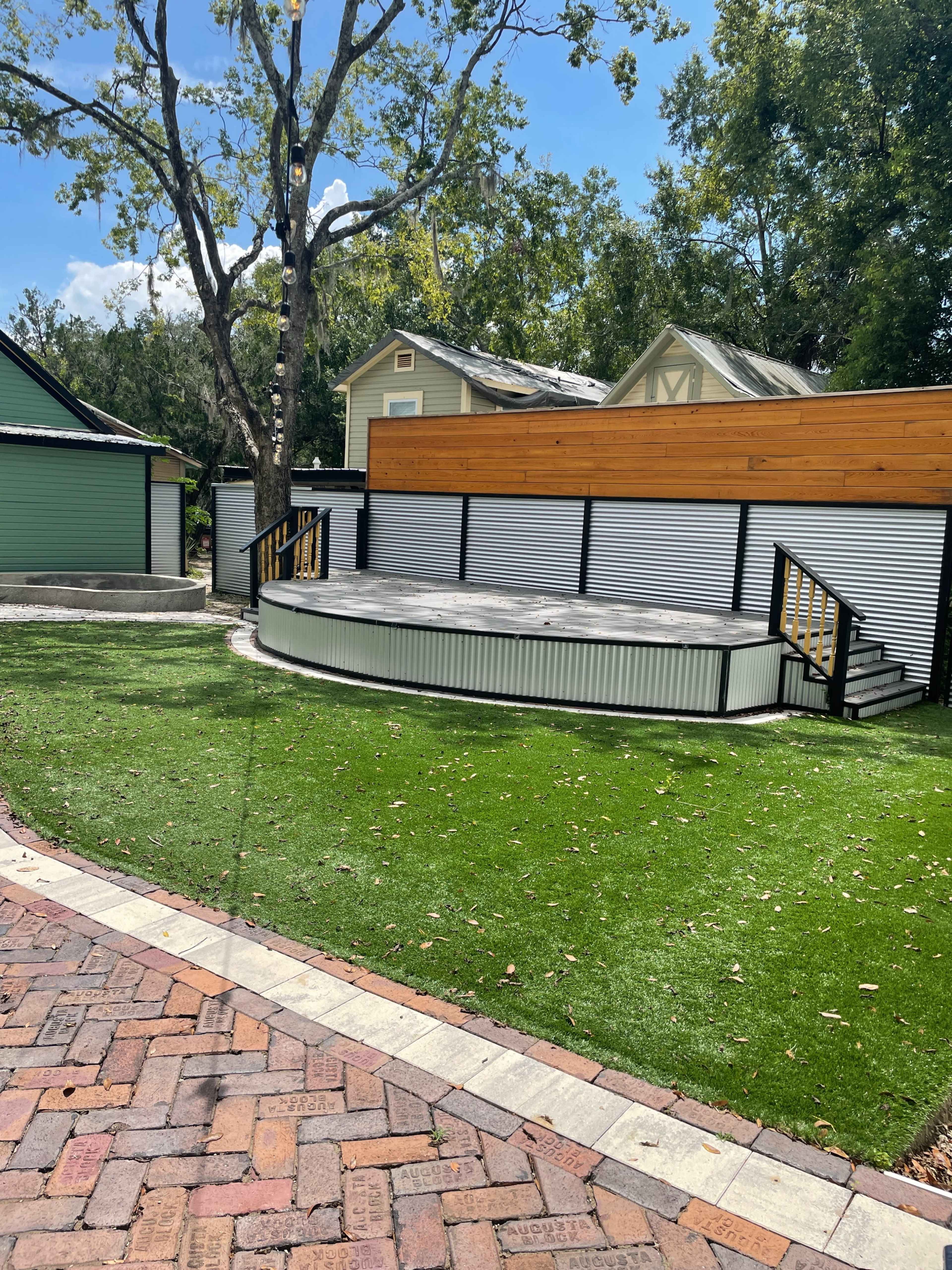 The image shows a circular wooden deck surrounded by artificial turf, located in a landscaped yard with nearby buildings.