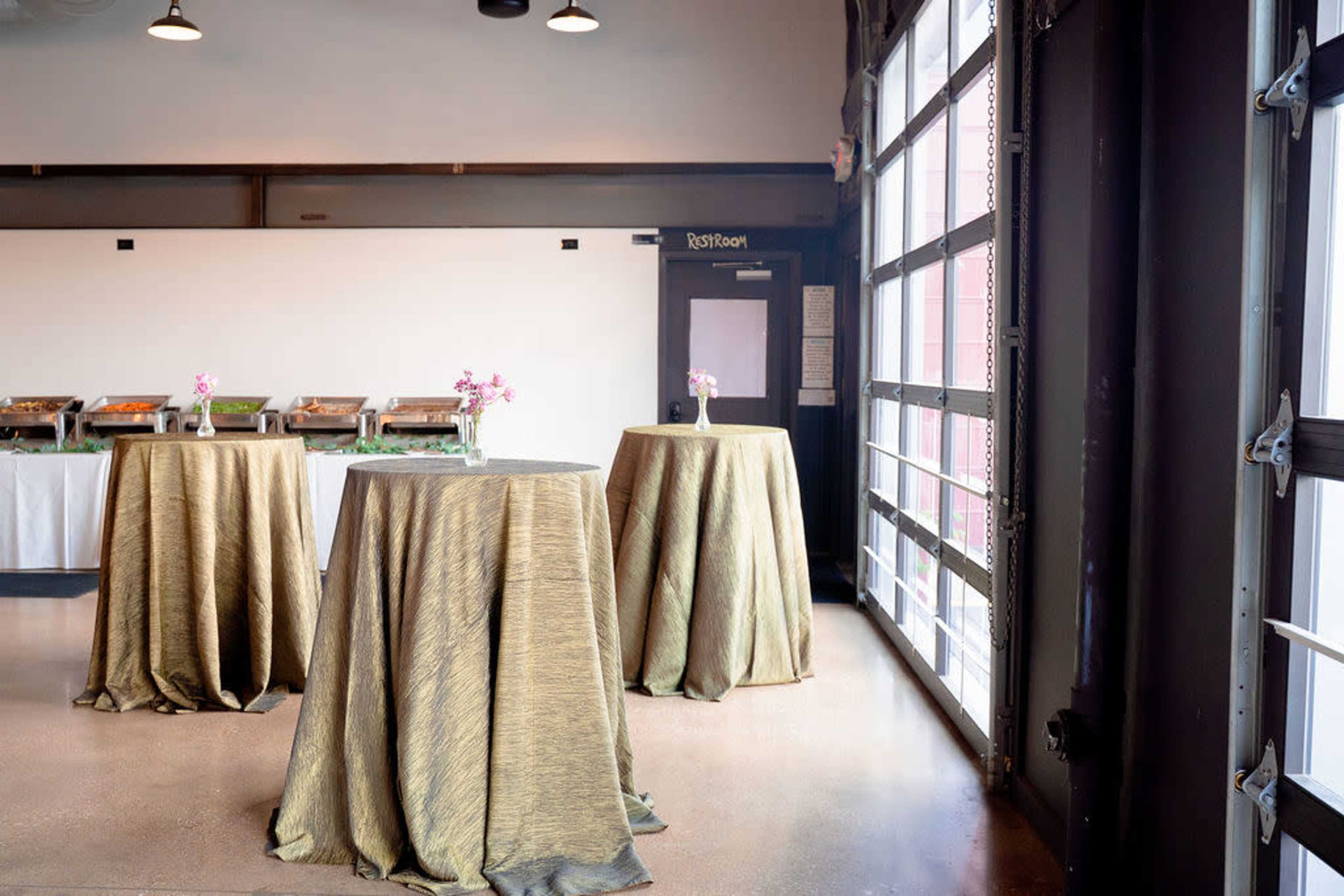 The image shows several high tables with gold-colored tablecloths set up in a room featuring a large window and a serving area in the background.