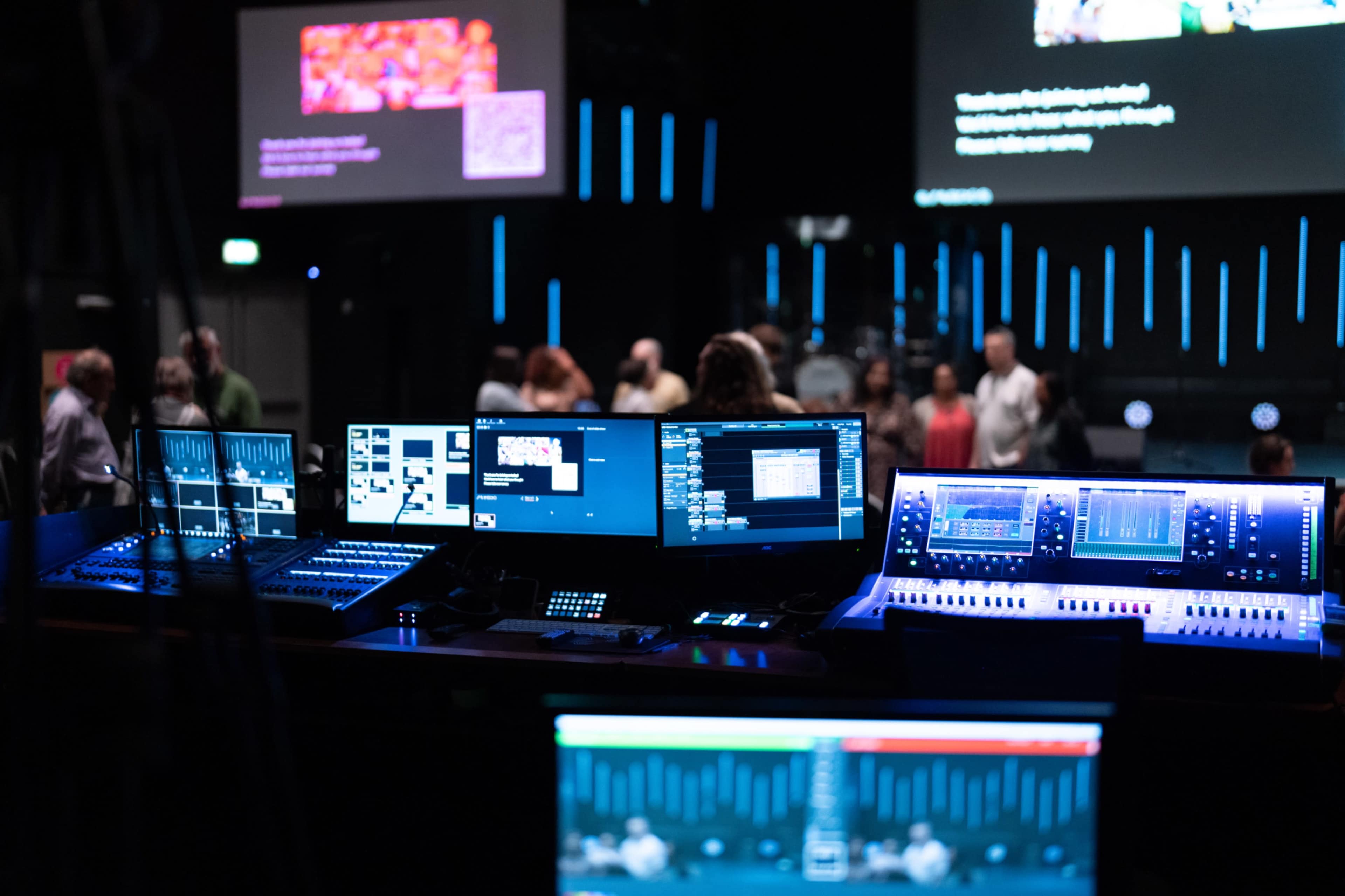 The image shows a sound and video control room with multiple screens displaying audio and visual settings, and people gathered in the background.