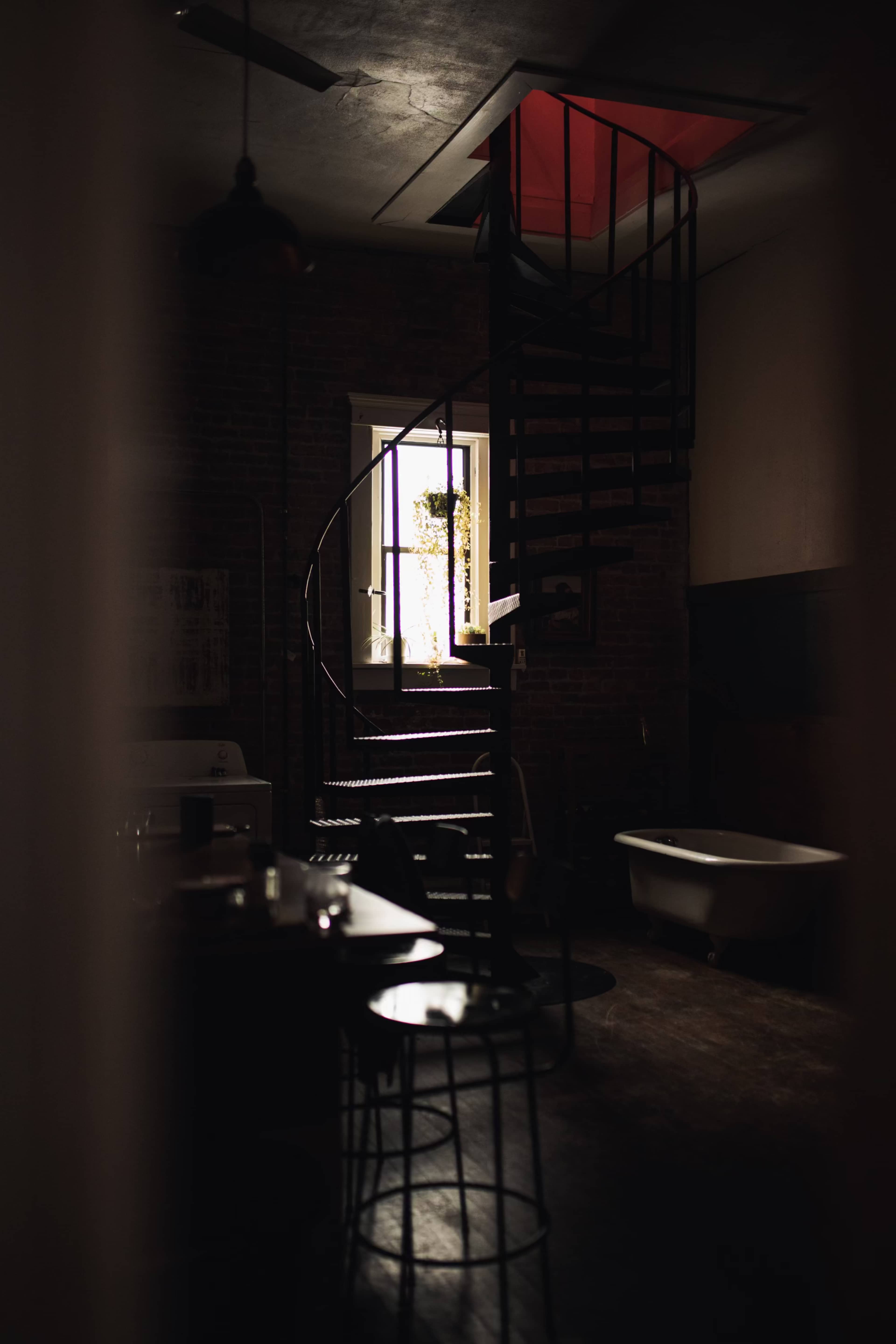 A spiral staircase leads up to a window with light streaming in, illuminating a dimly lit room with a bathtub and a small table.