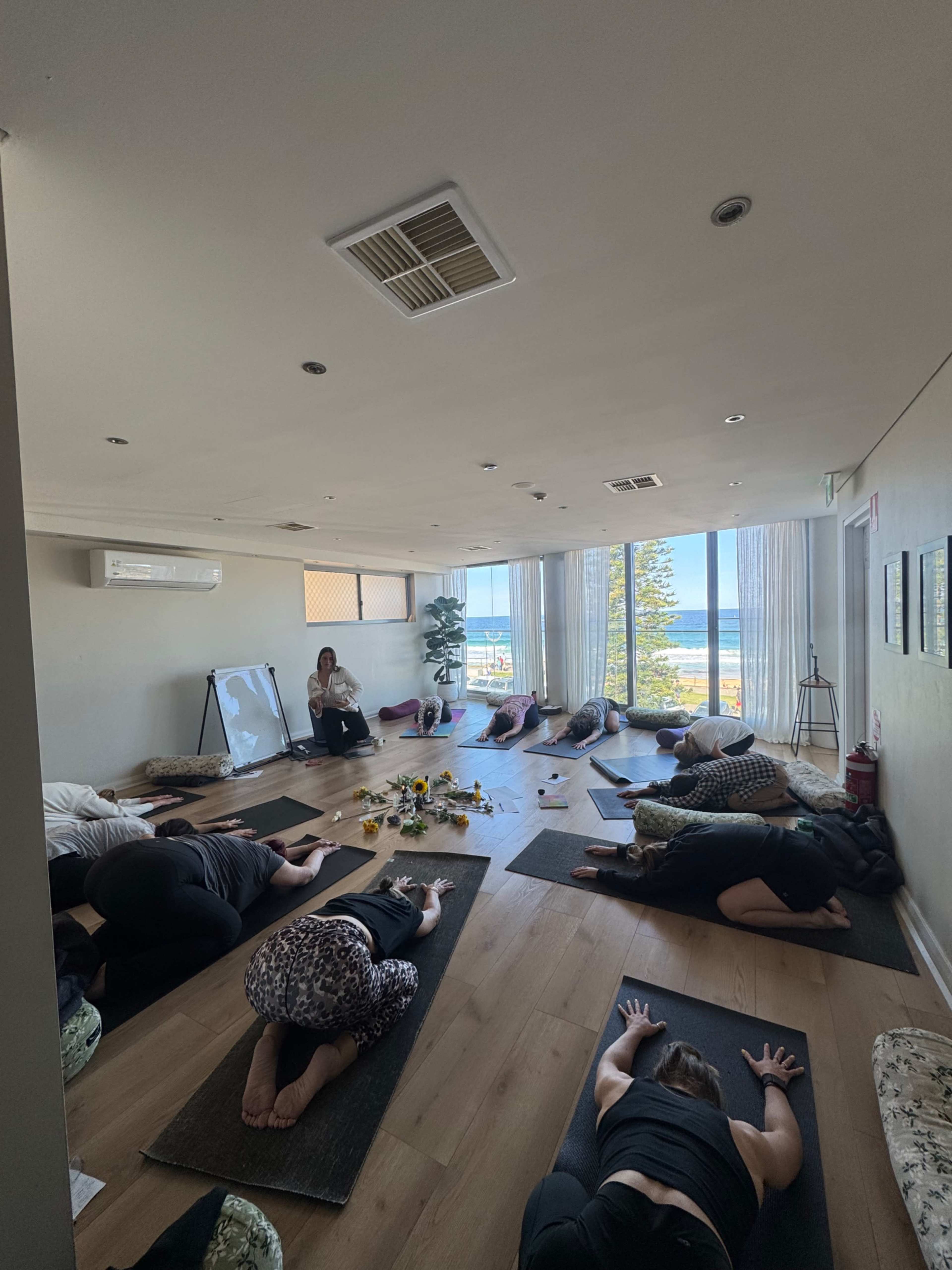 A group of people is practicing yoga in a studio with large windows overlooking a scenic view of the ocean.