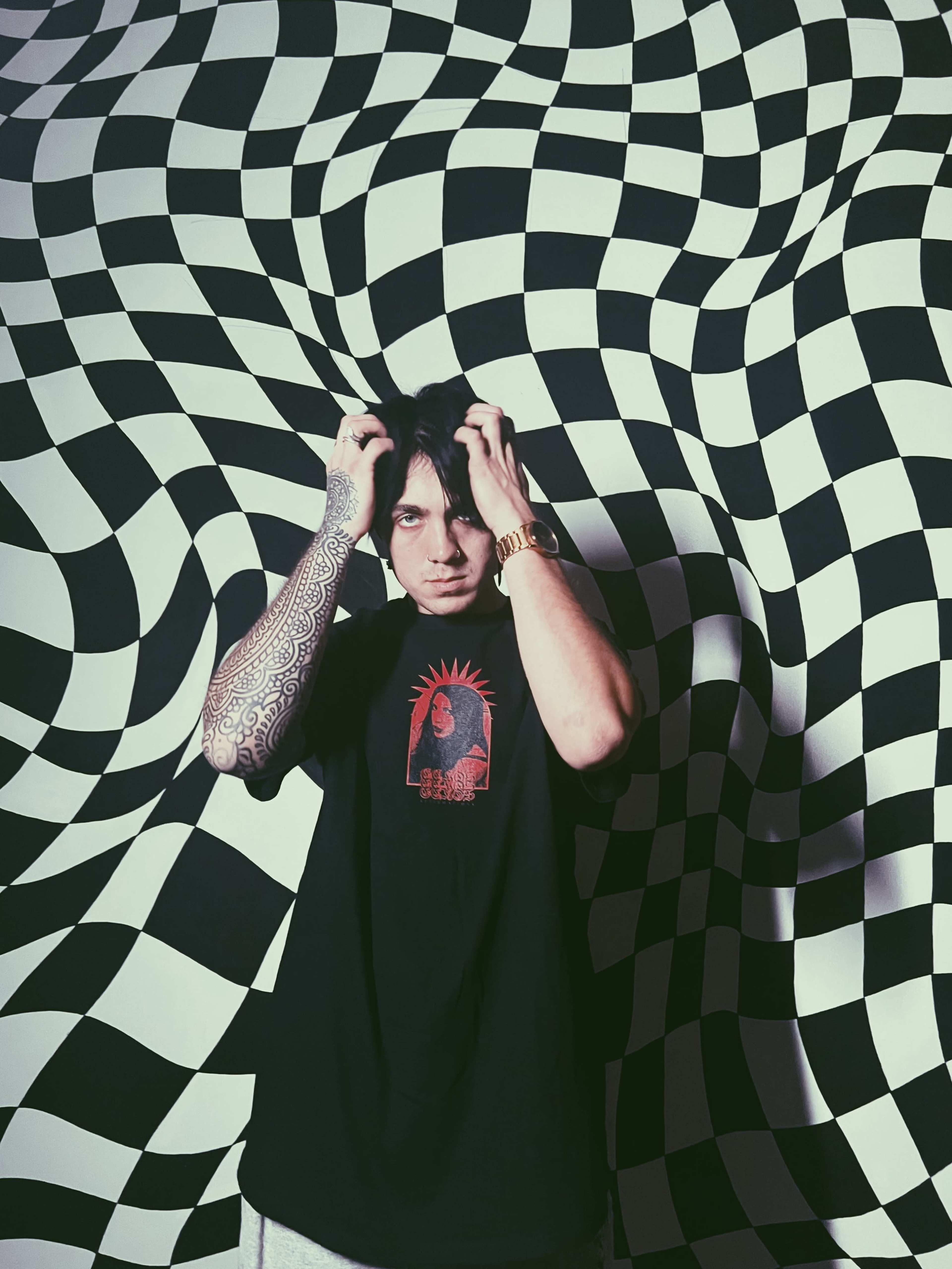 A person with a tattooed arm stands in front of a black and white checkered wall, holding their head in their hands.