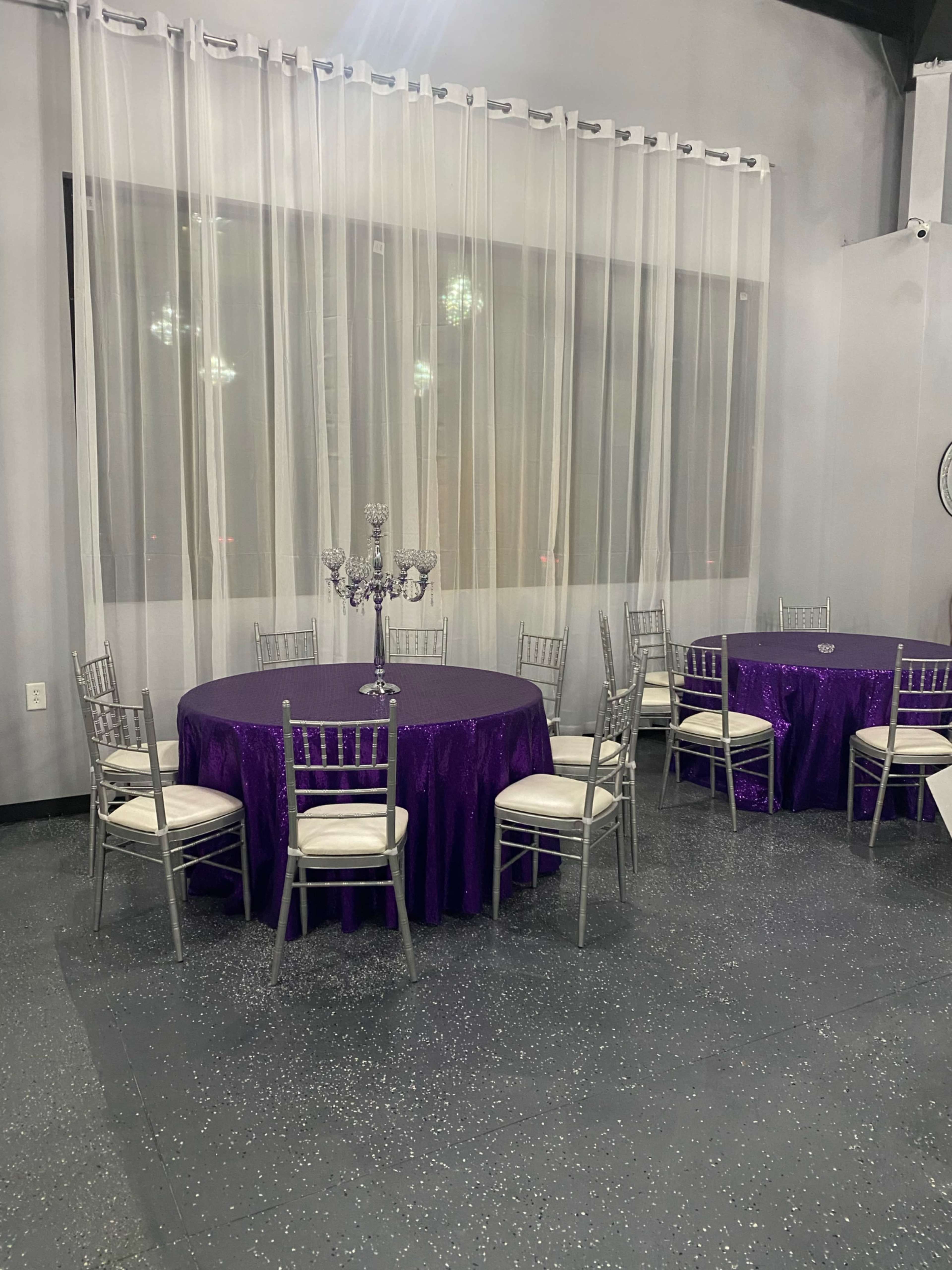 A banquet room with round tables covered in purple tablecloths surrounded by silver chairs, set against a backdrop of sheer curtains.