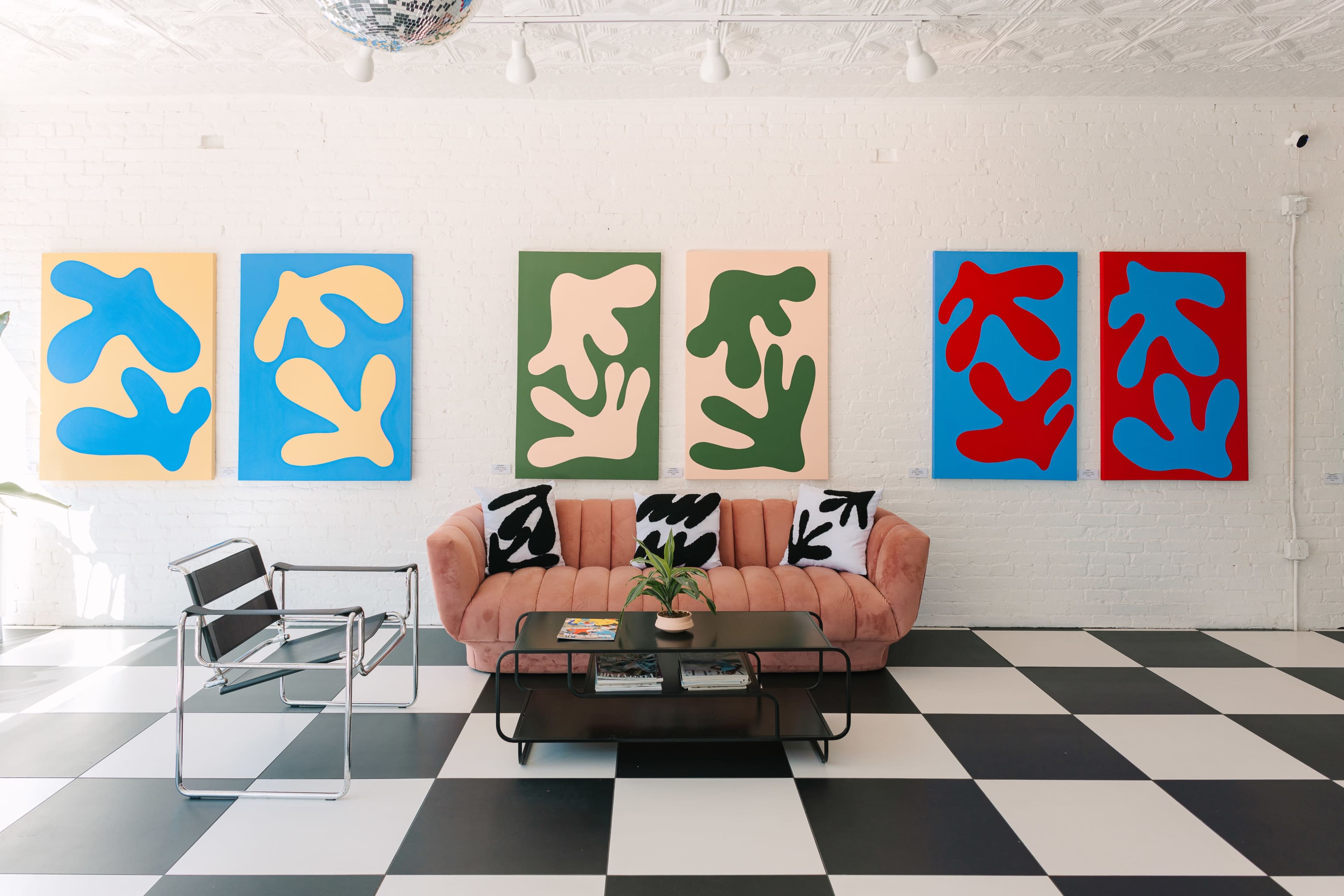 A pink sofa is placed in a room with black and white checkered flooring, featuring large abstract paintings on the white walls and a glass coffee table with a plant on it.