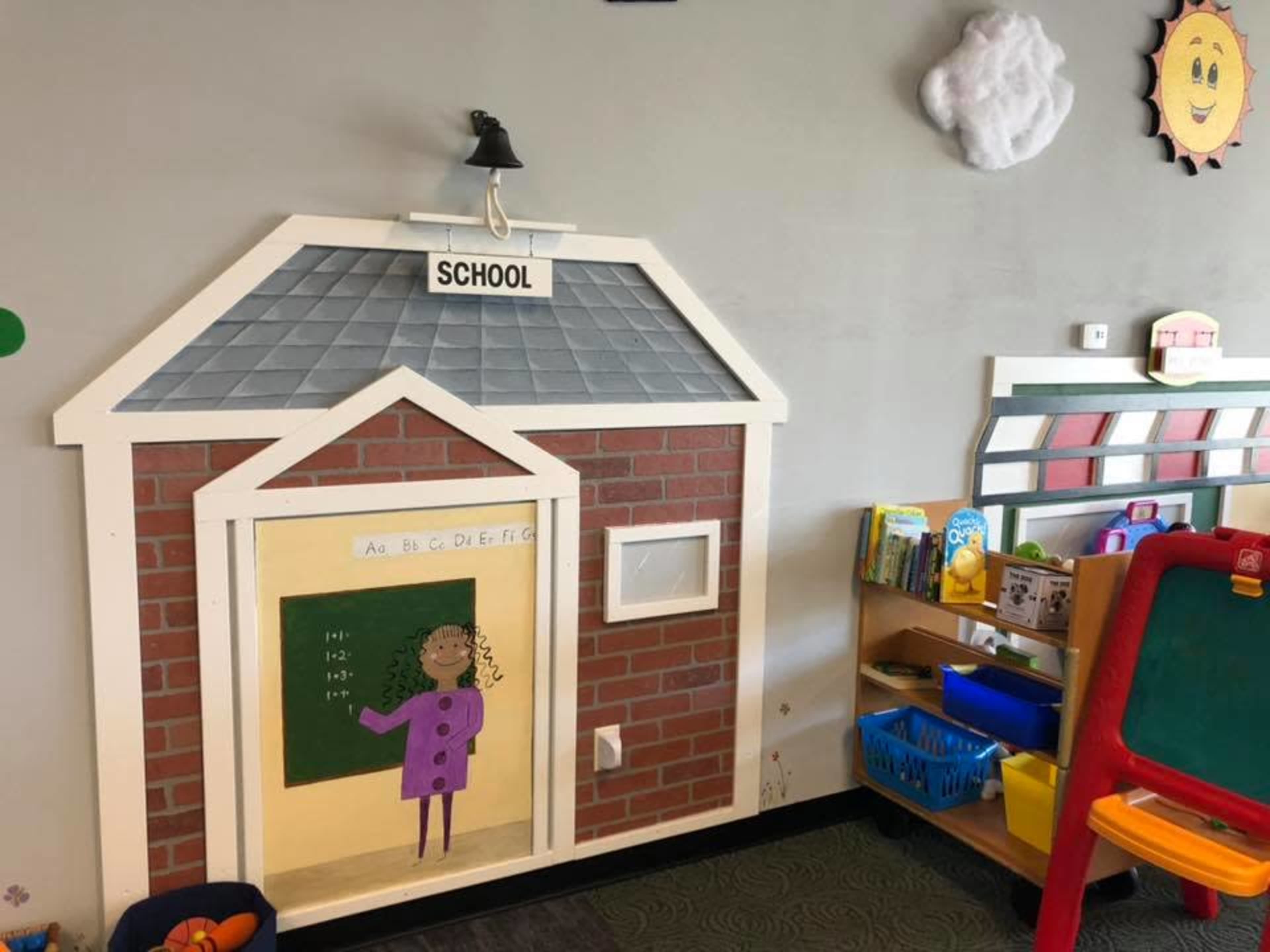 A classroom corner with a painted wooden structure resembling a schoolhouse, complete with a chalkboard at the entrance and colorful educational materials on shelves nearby.