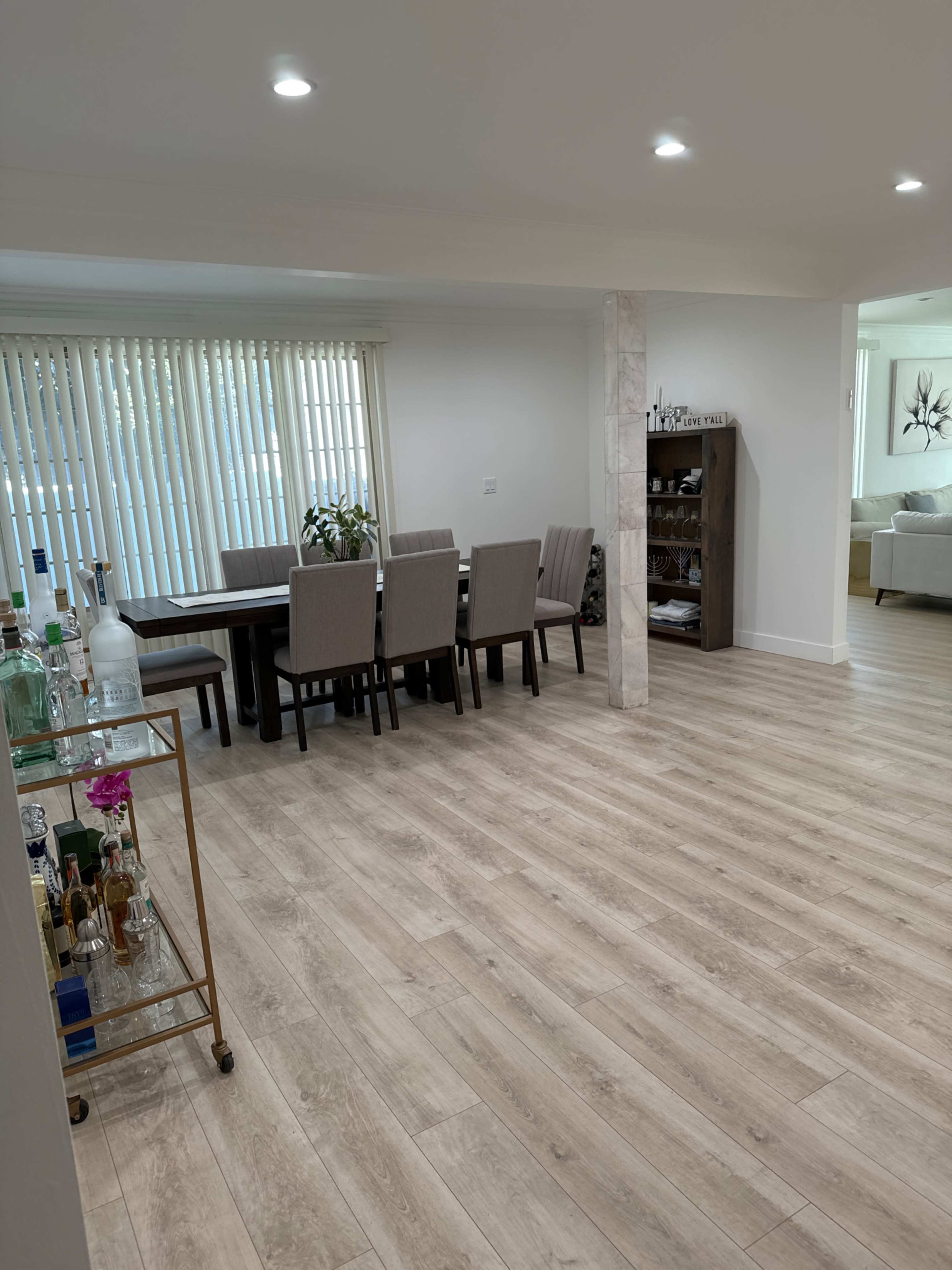 A modern dining area with a table and six chairs, a bar cart on the left, and a bookshelf against the wall.