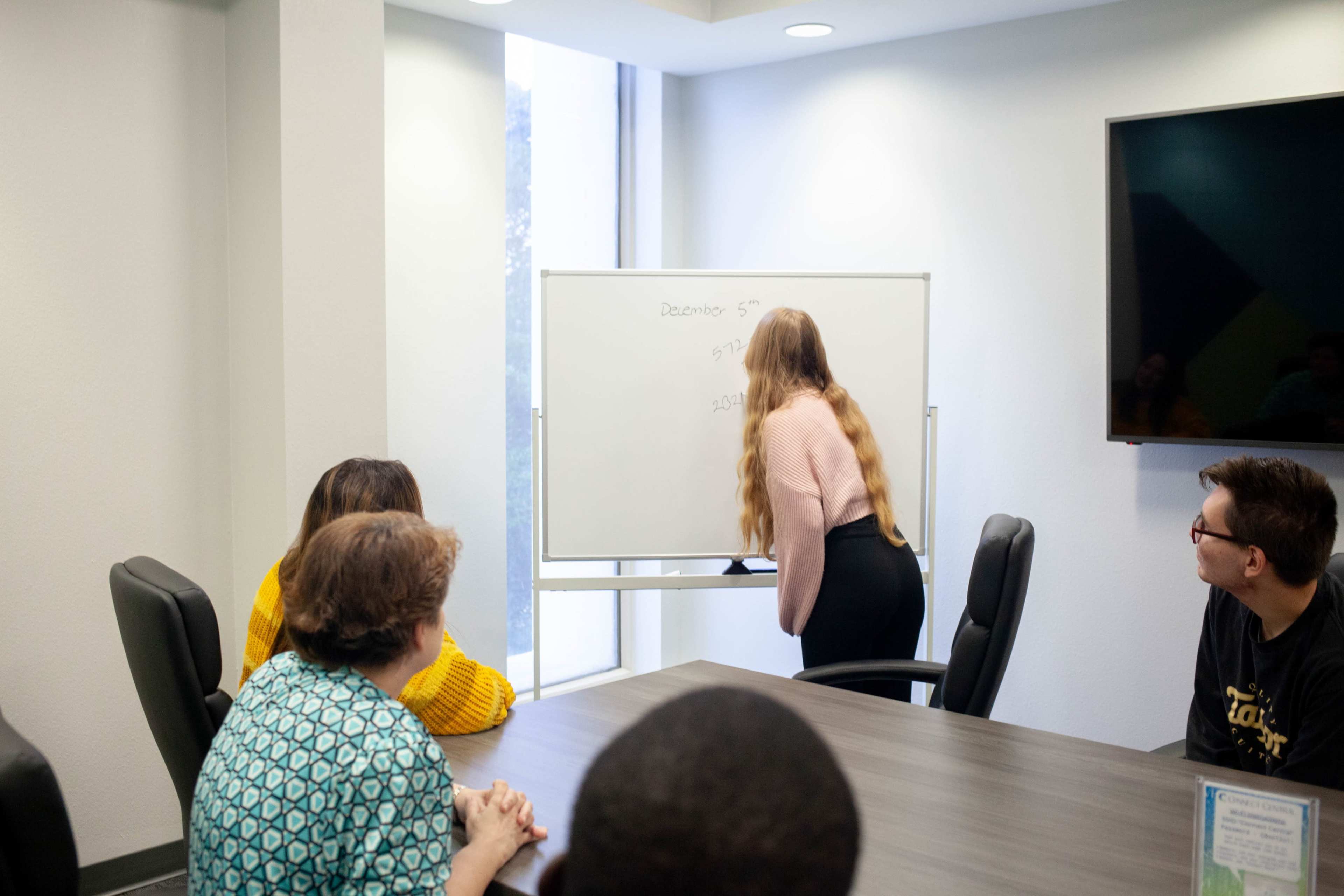 A group of people watches as a person writes on a whiteboard in a conference room.