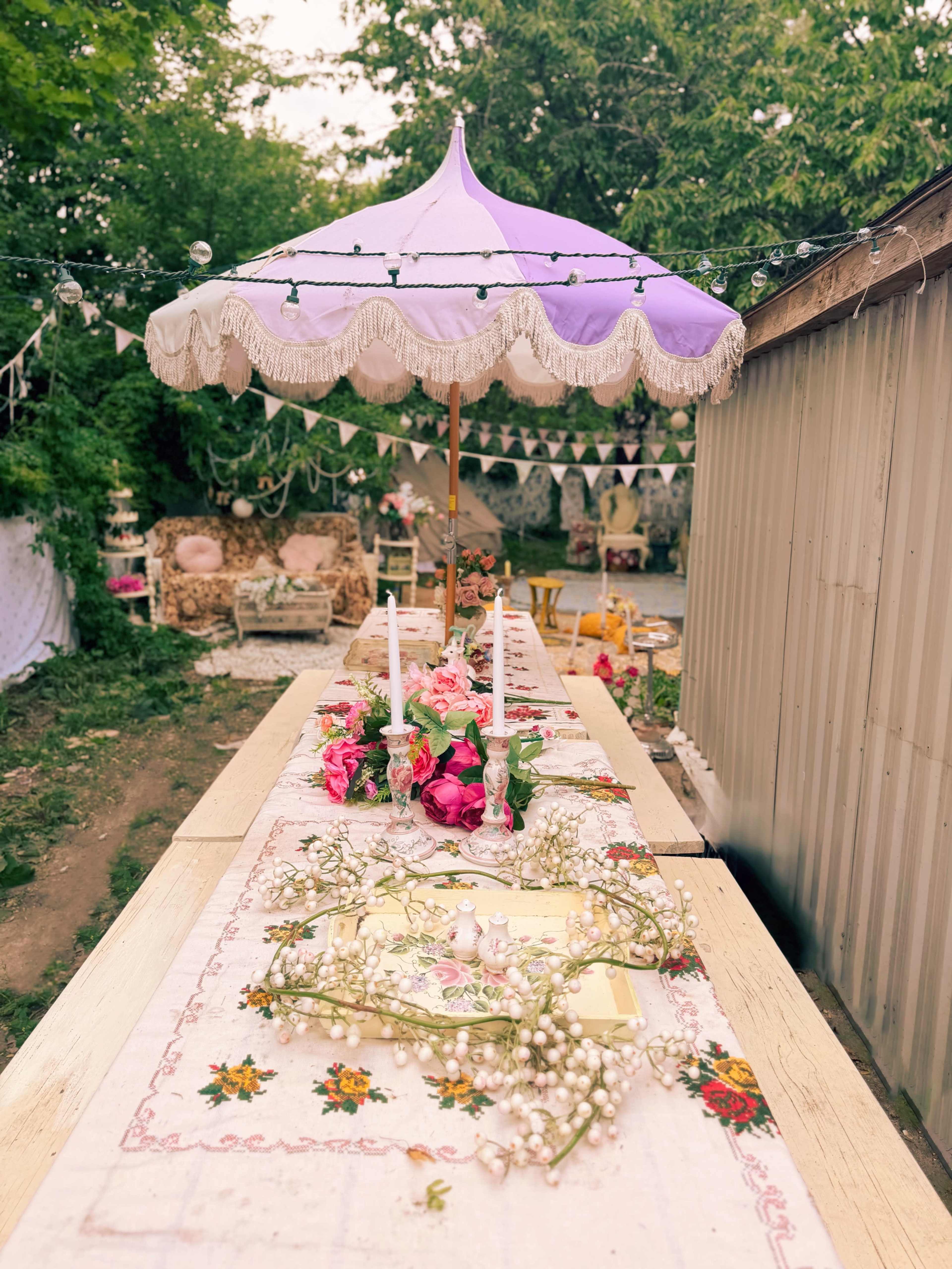 A long outdoor table is set with floral arrangements and candles under a decorative purple umbrella, surrounded by a whimsical garden setting.