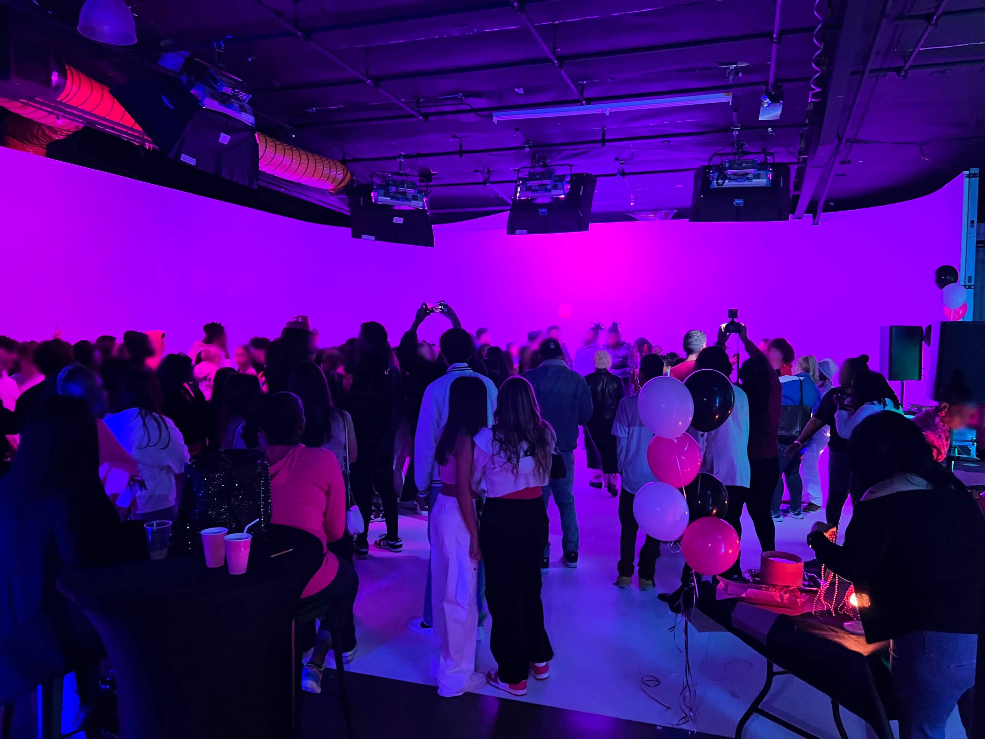 A crowd of people gathers in a dimly lit space with purple lighting, enjoying an event with tables set up and music playing.