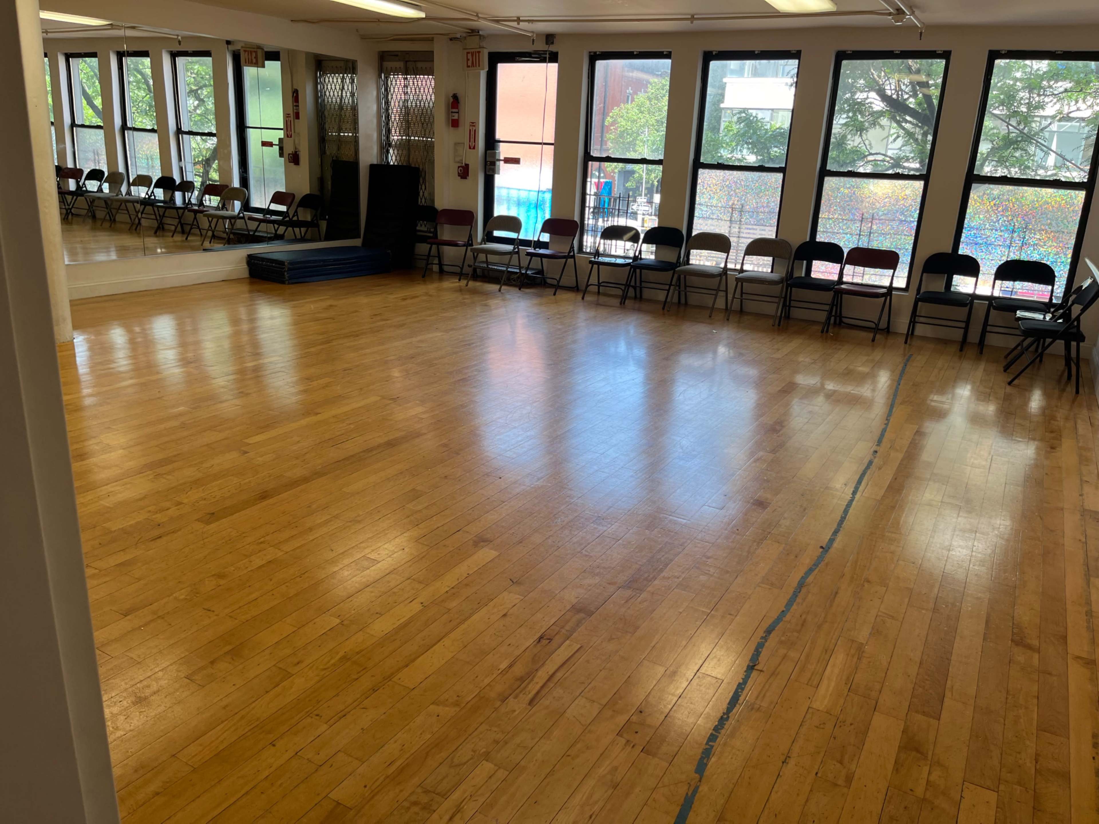A spacious dance studio with wooden floors, lined with chairs along the walls and large windows allowing natural light.