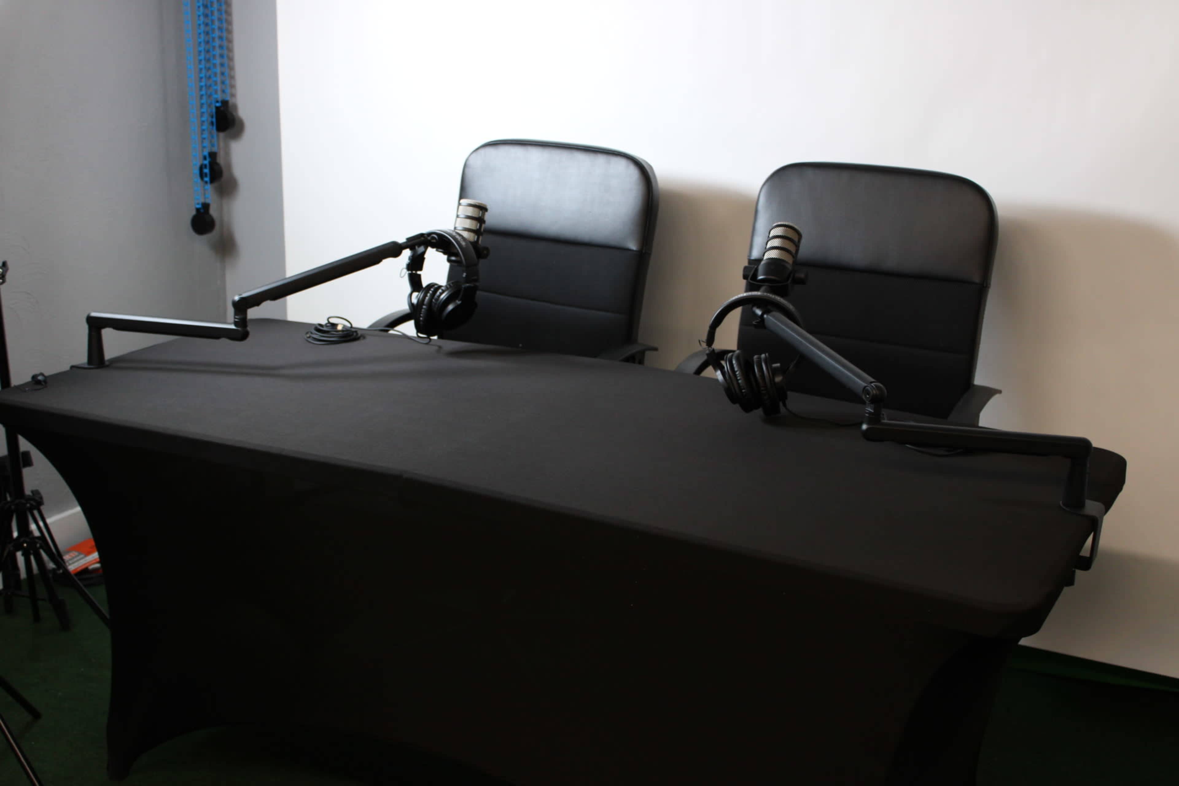 The image shows a black table with two black chairs and mounted microphones, set up for a podcast recording.