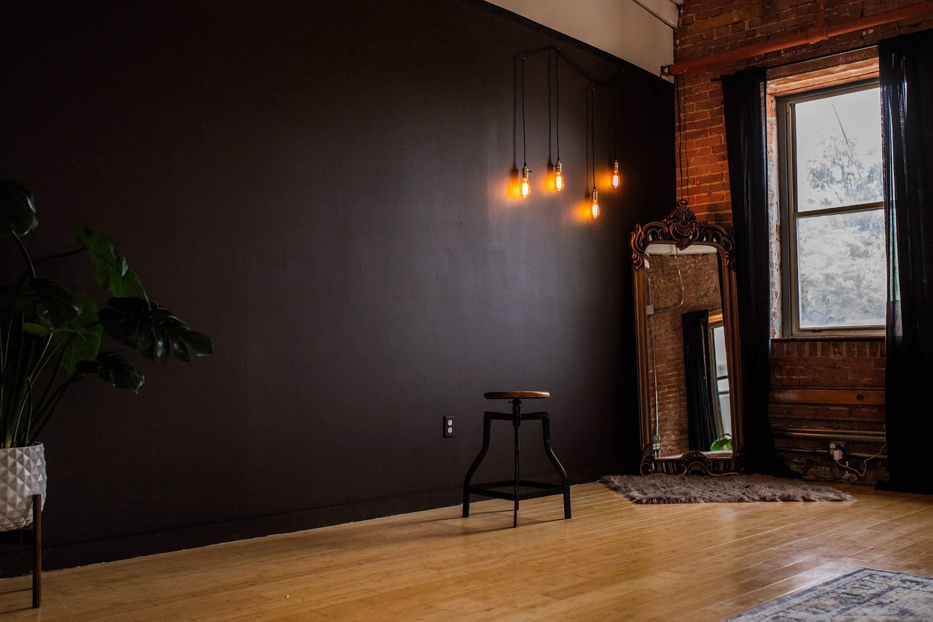 The image shows a minimalist interior space featuring a dark wall, a wooden floor, a small stool, a mirror, and a window with flowing curtains.