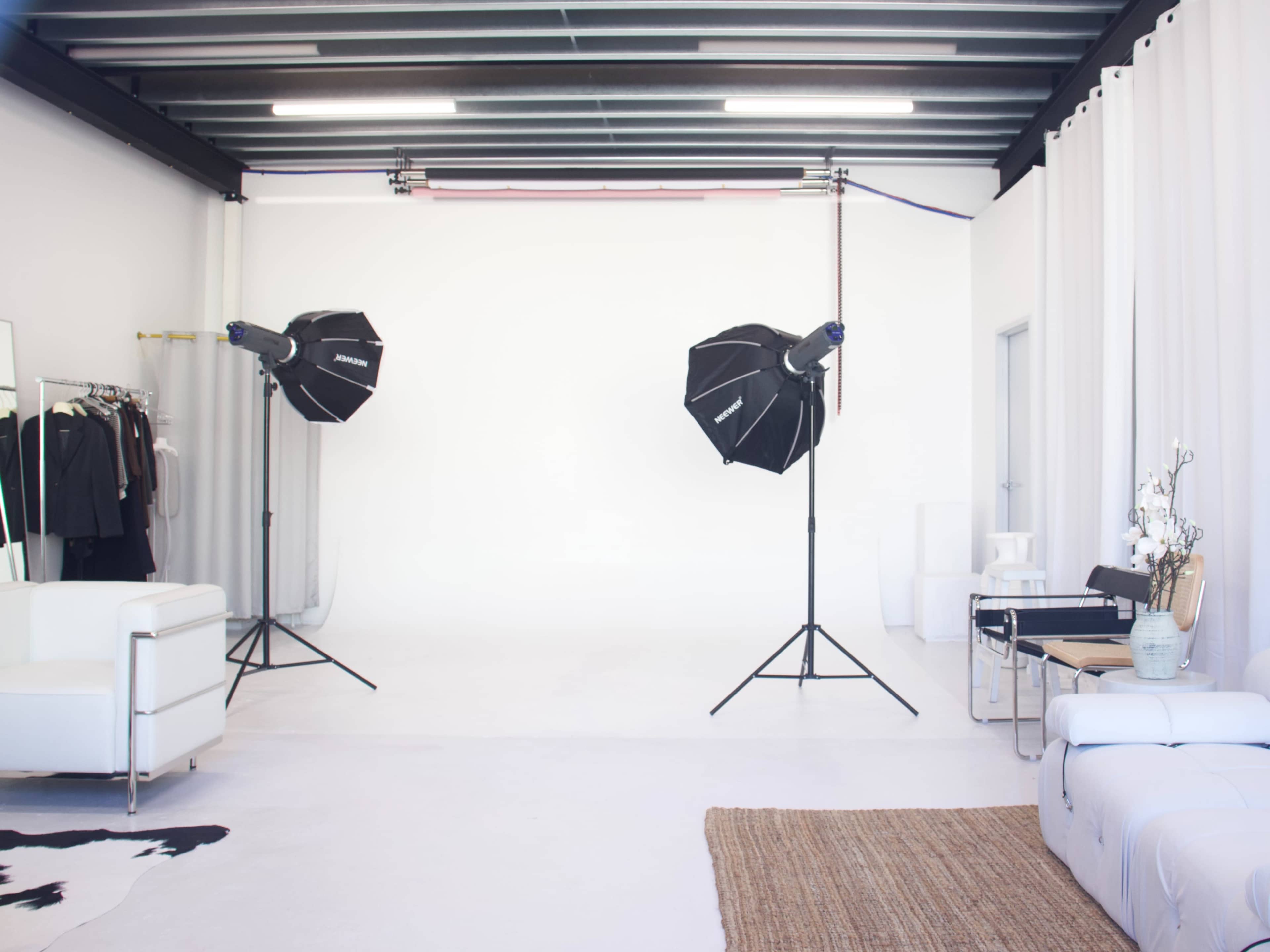 The image shows a photography studio with two light stands, a white backdrop, and various props and furniture arranged in the space.