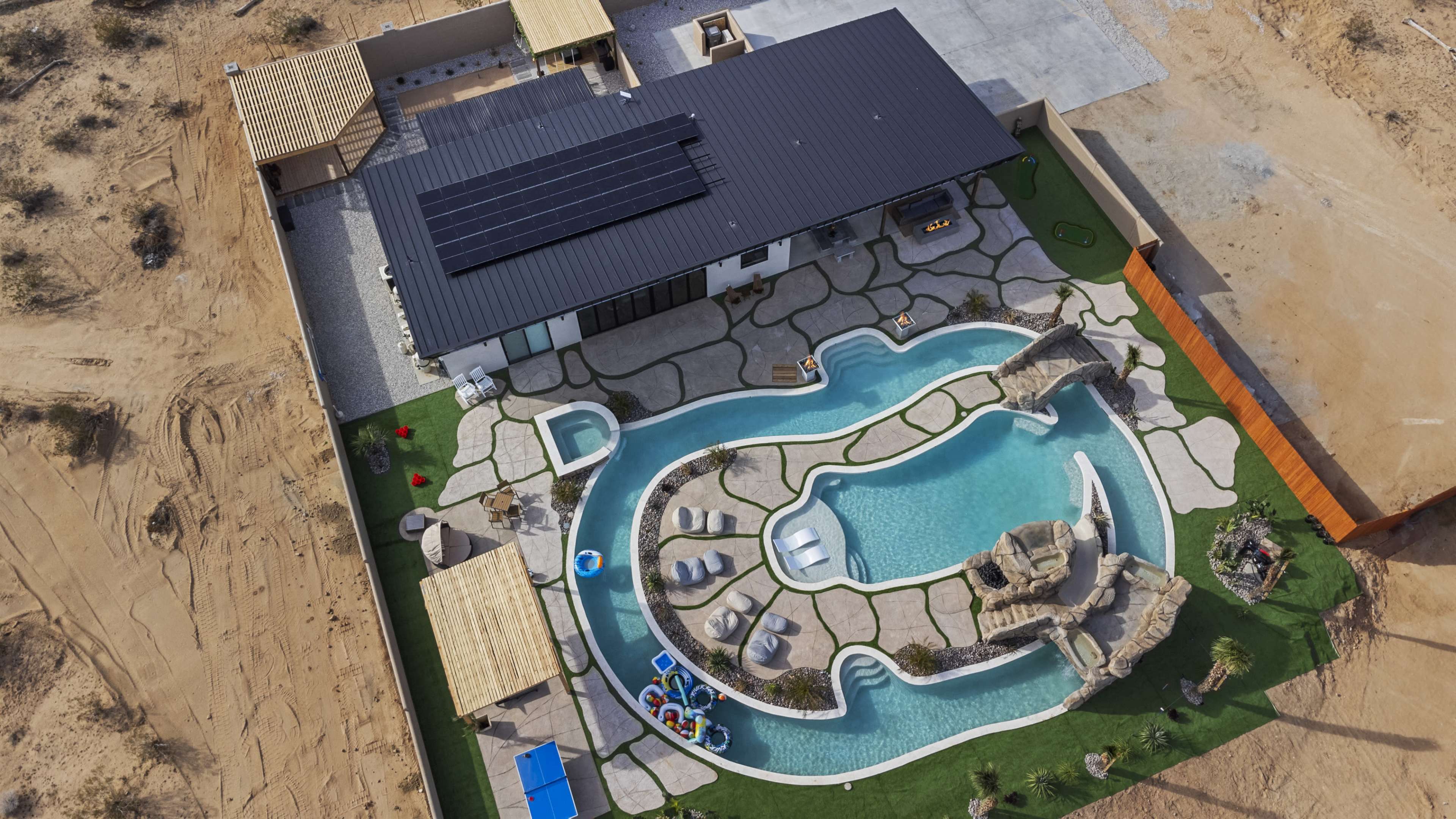An aerial view of a modern house with a landscaped backyard featuring a winding pool, rock formations, and lounge areas.
