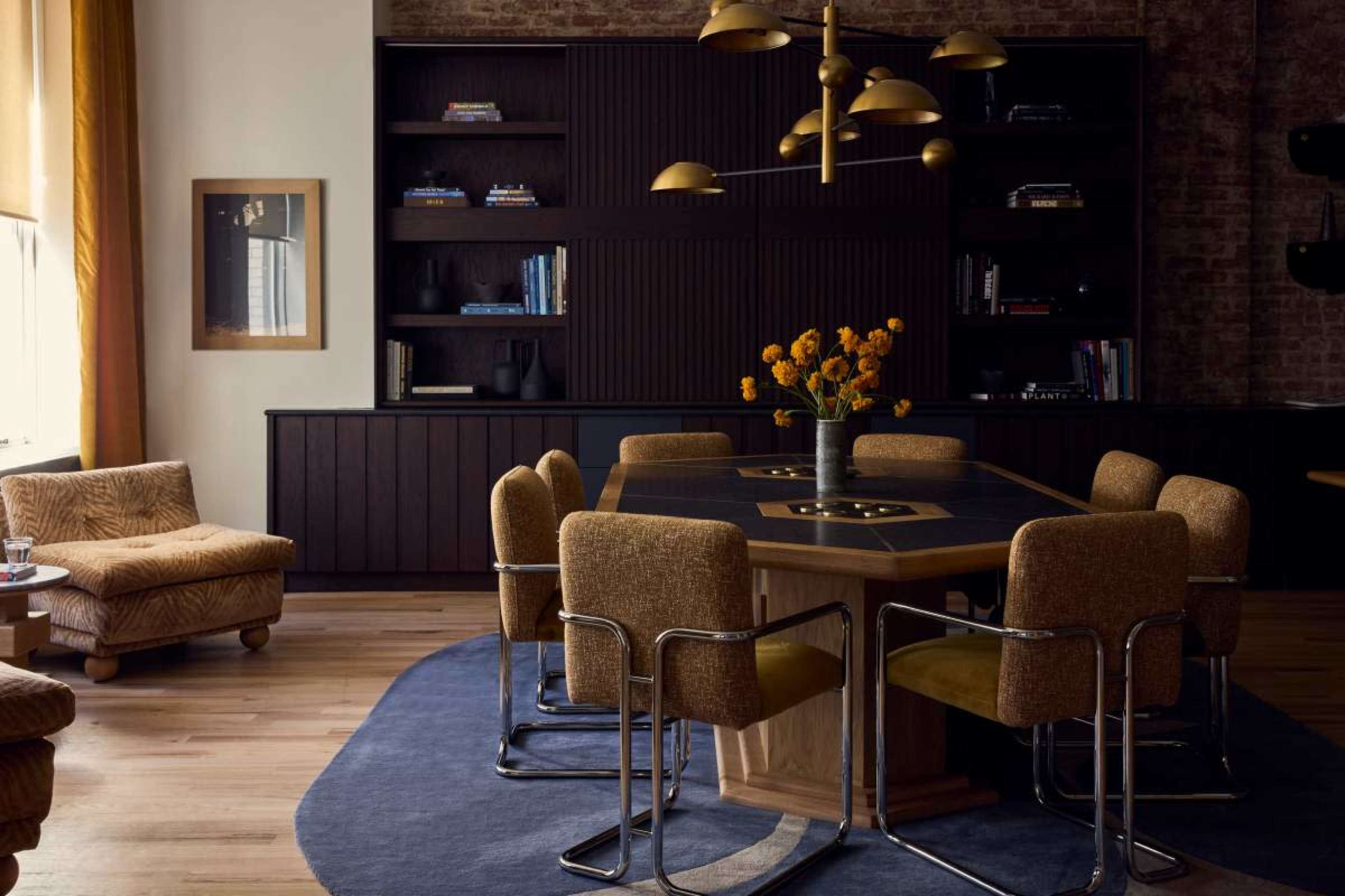 A modern dining room features a large wooden table surrounded by upholstered chairs, with a flower vase at the center and bookshelves in the background.