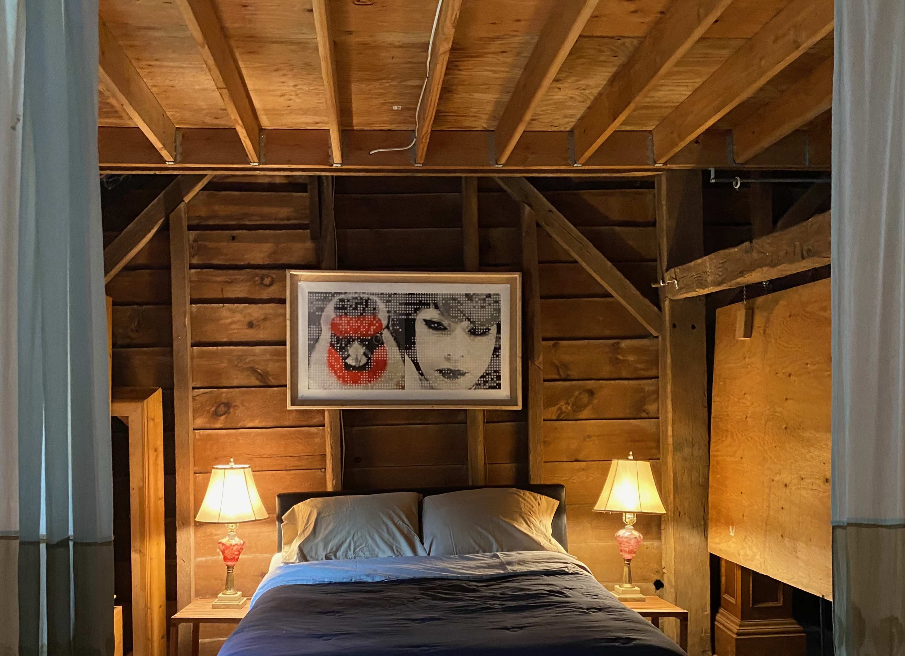 A bedroom features a bed with blue bedding framed by wooden walls and two bedside lamps, while a framed artwork hangs above the headboard.