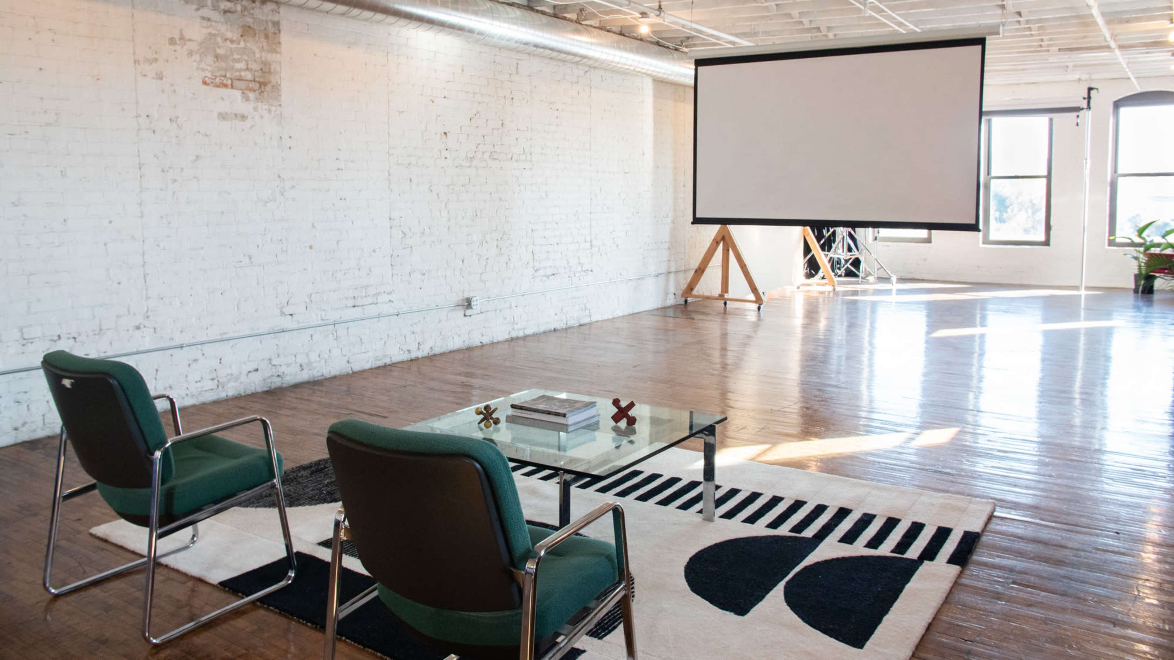 A spacious room with white brick walls, hardwood floors, a large projection screen, a glass coffee table, and two green chairs on a patterned rug.