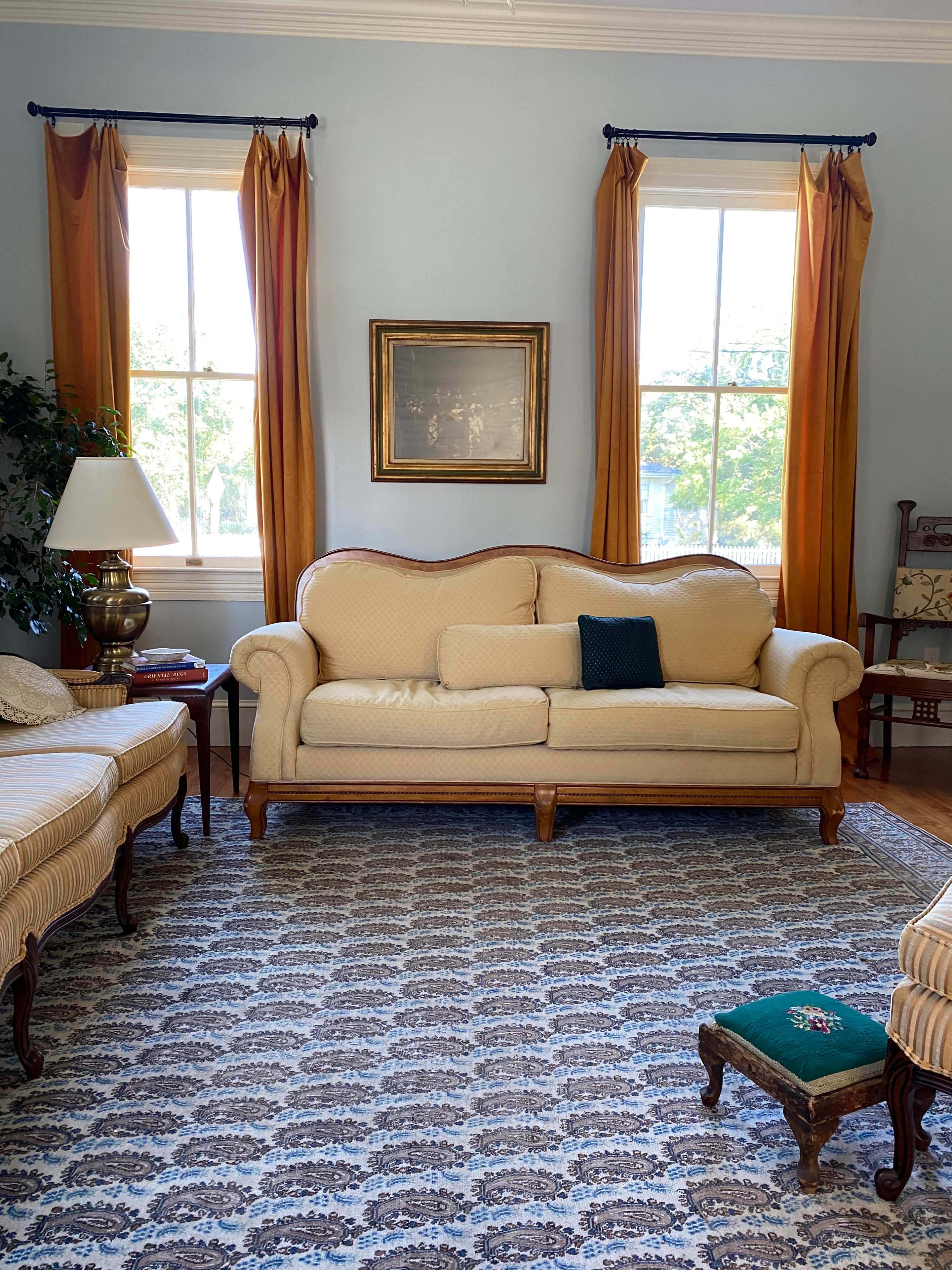A light-colored living room with a sofa, decorative pillows, a lamp, and large windows draped with golden curtains.