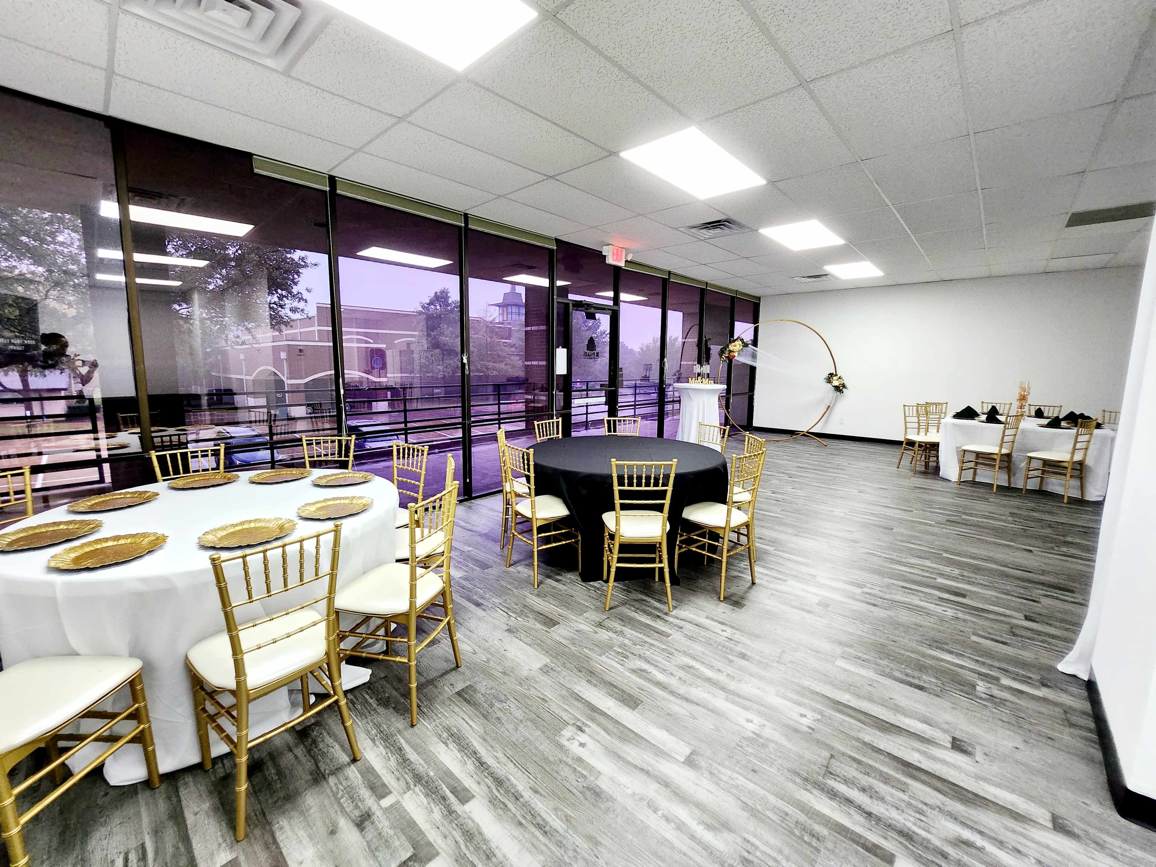 An event space with multiple tables set up, including a black table and a white table, all surrounded by gold chiavari chairs, under bright overhead lights.