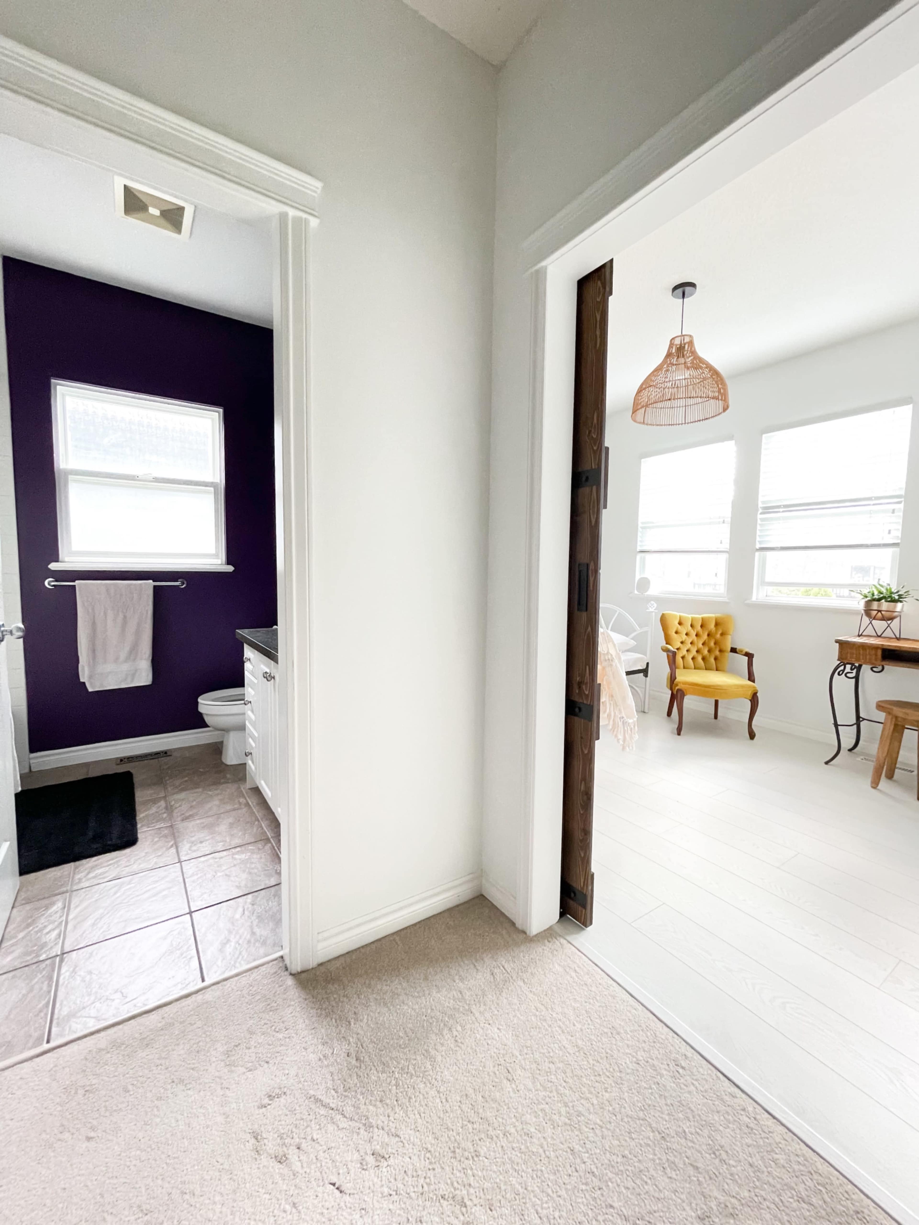 The image shows a hallway leading to a bathroom with a purple accent wall and a room with large windows and a yellow chair in the background.