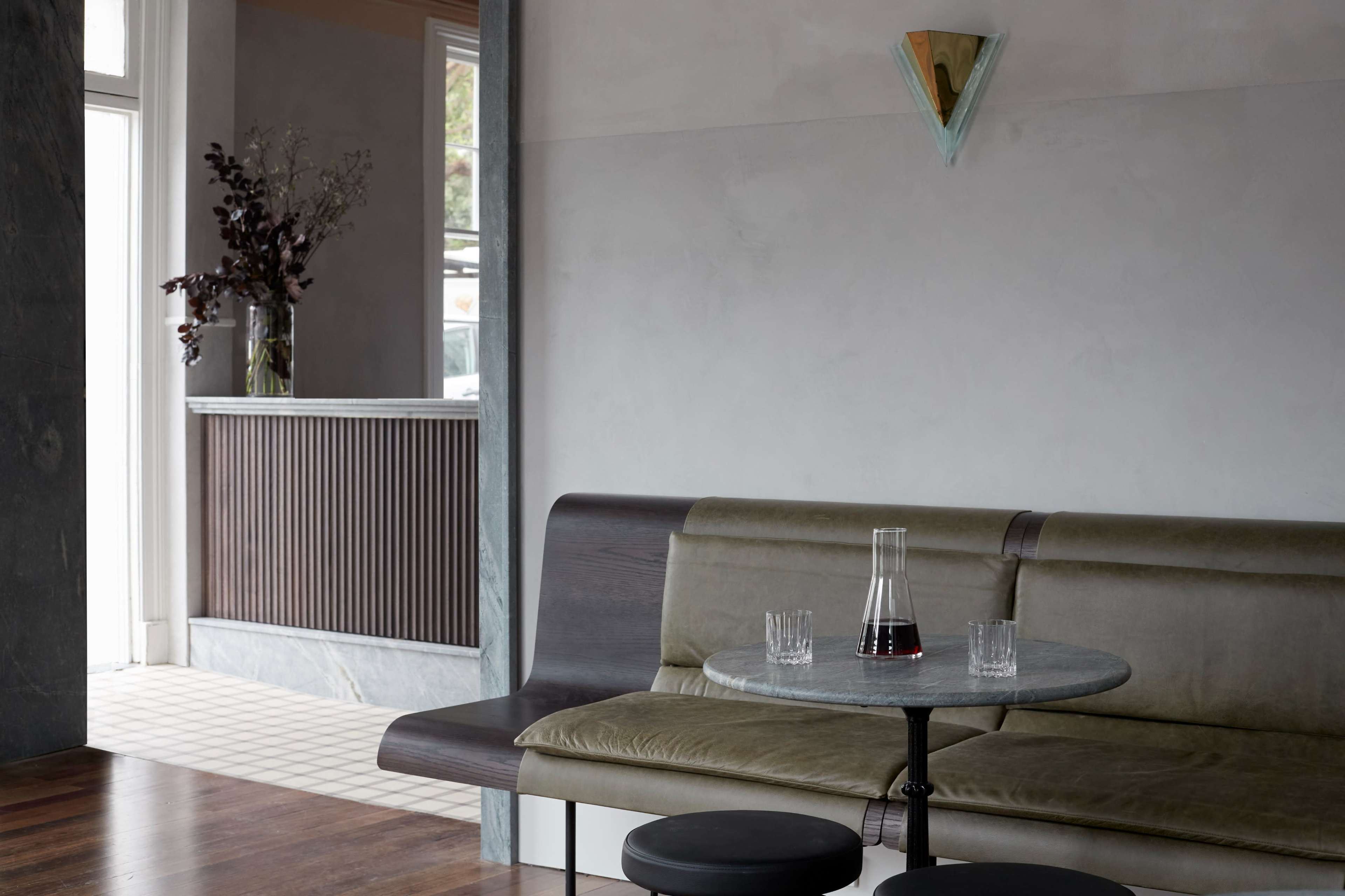 A minimalistic interior features a grey wall, a dark wooden bench with a round table, and a reception area with a marble counter in the background.