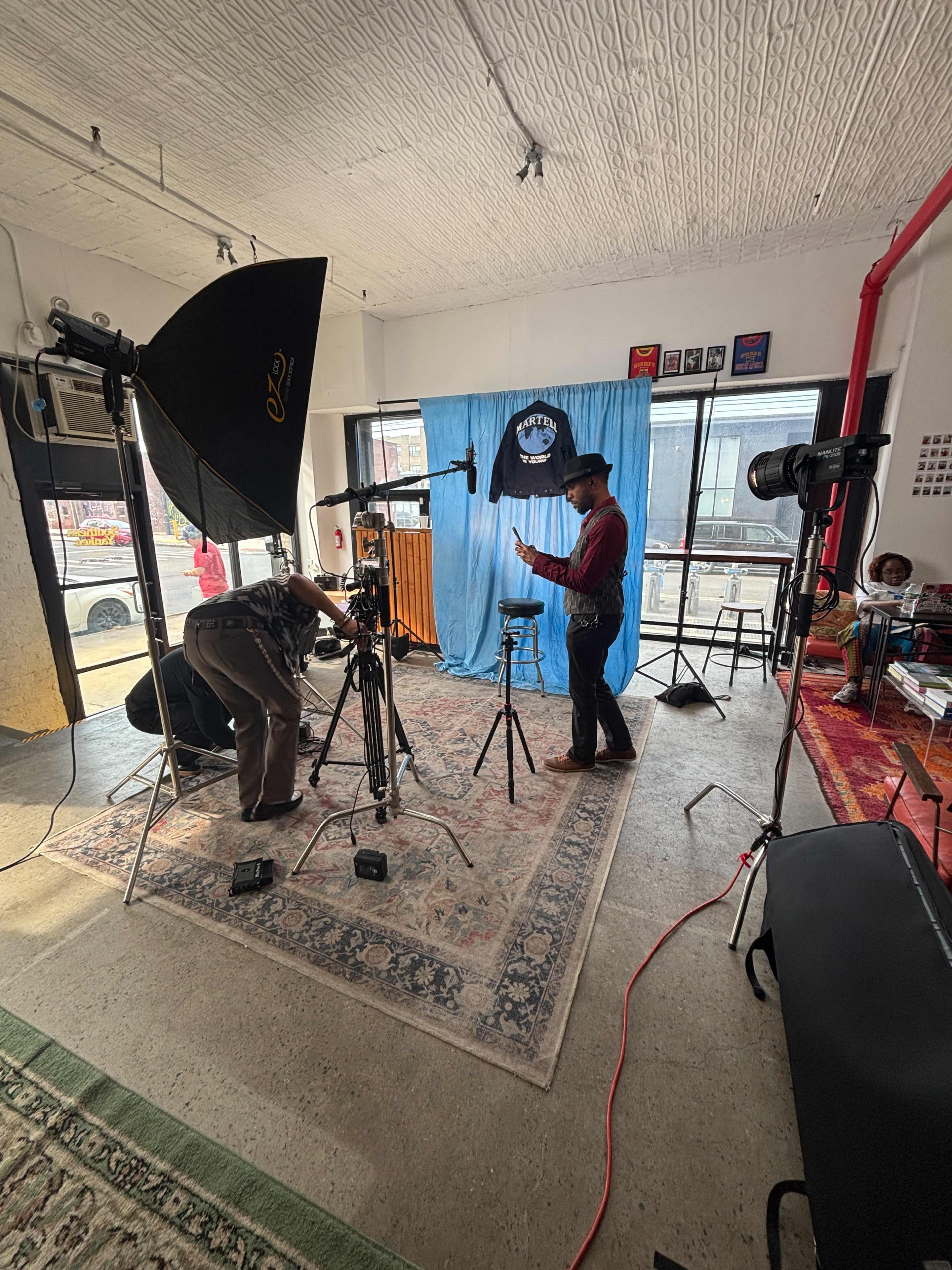 A film crew sets up equipment in a studio featuring a blue backdrop, with one person preparing a camera while another adjusts lighting.