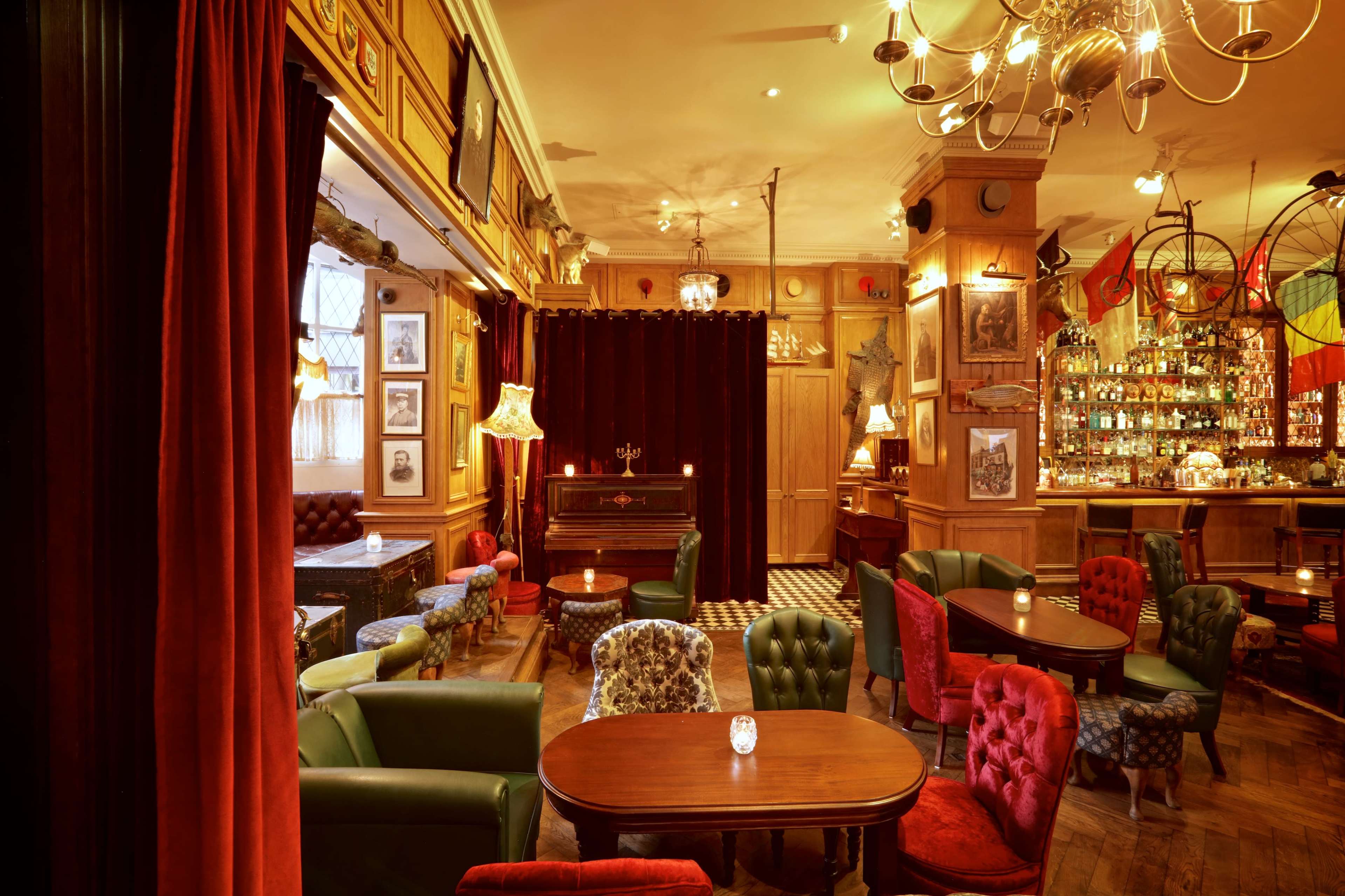 The image depicts a cozy bar with vintage furnishings, including green and red upholstered chairs, wooden tables, and a softly lit interior adorned with framed pictures and decorative elements.