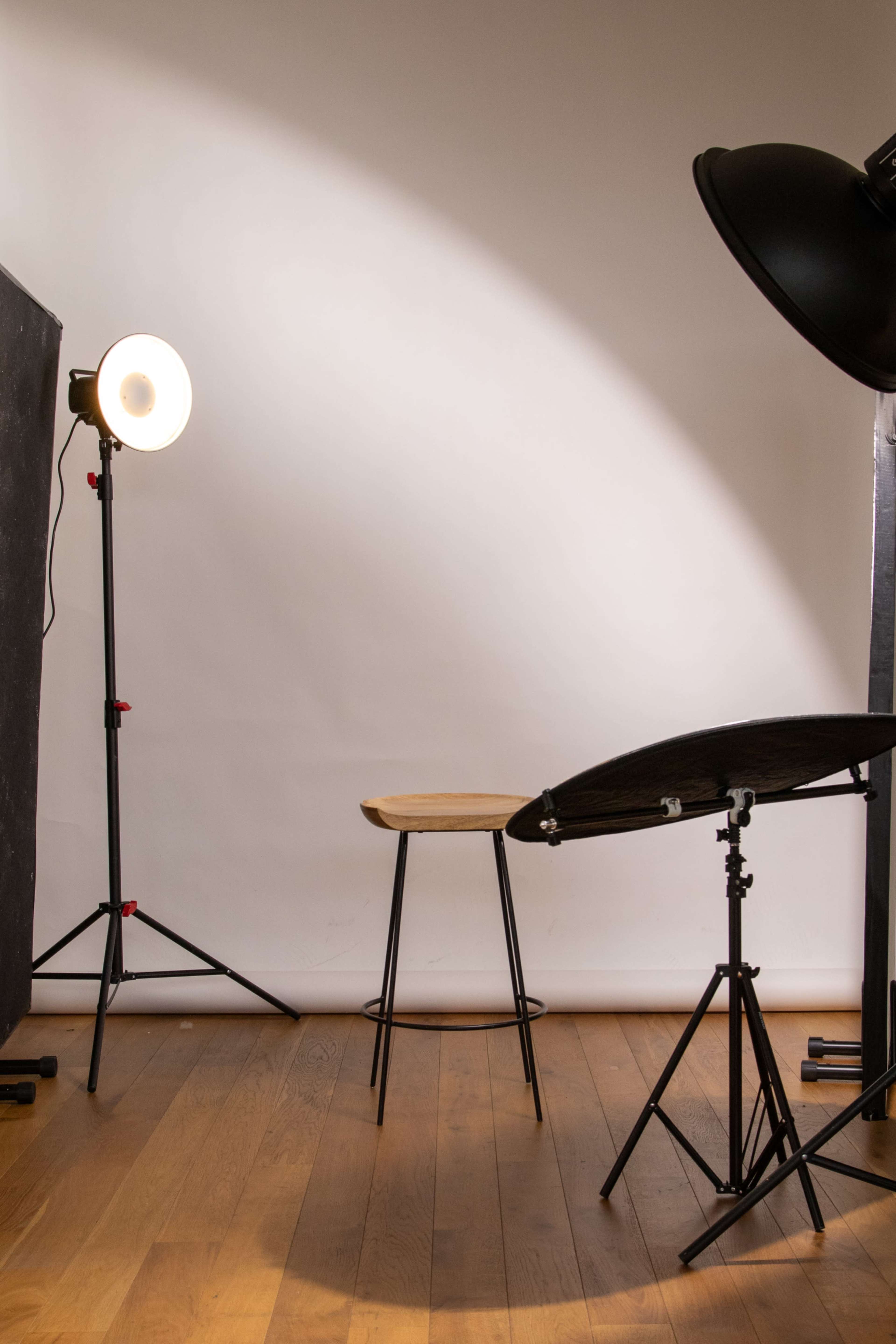 A wooden stool is positioned in front of a plain white backdrop, surrounded by studio lighting equipment.