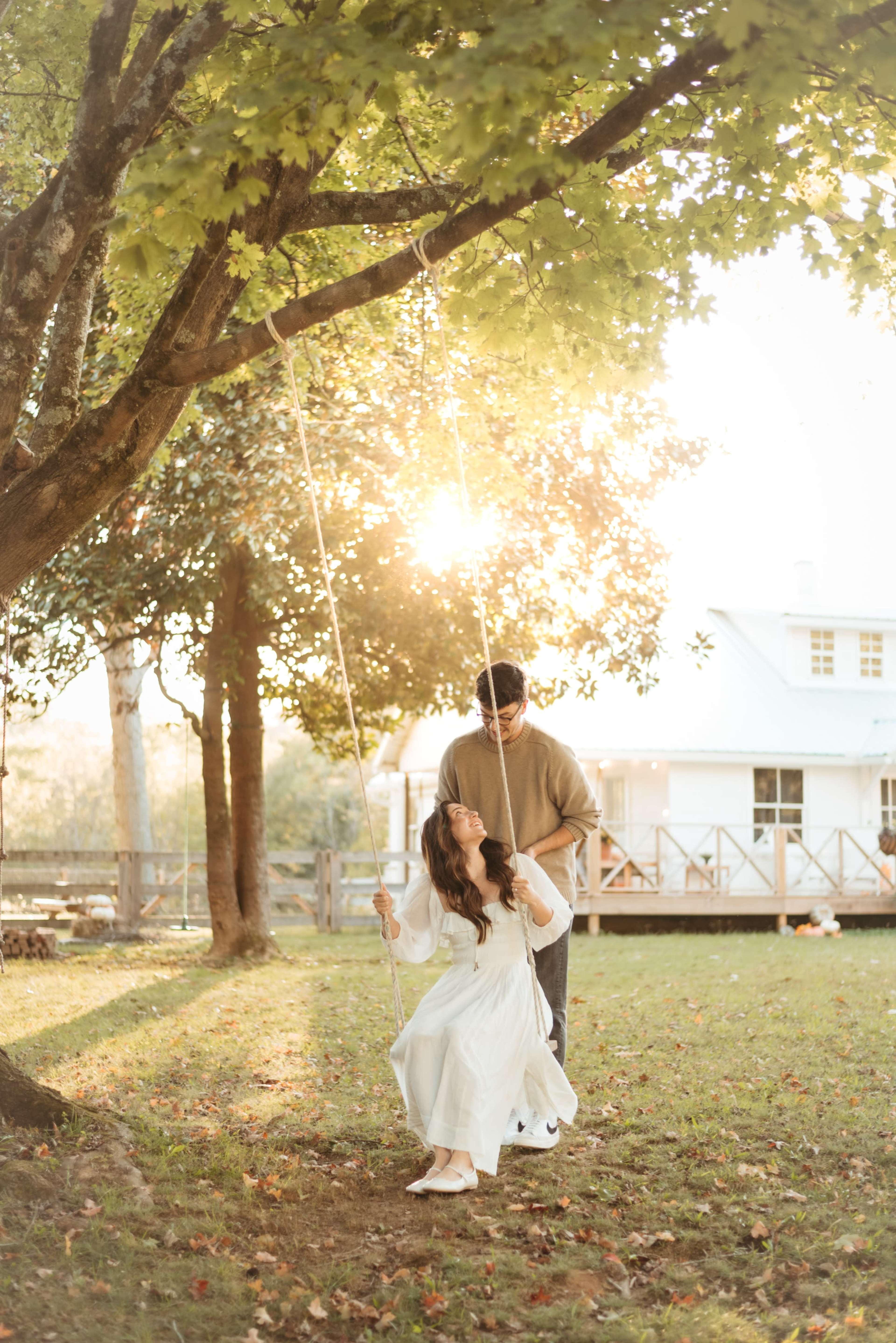 A woman in a white dress swings under a tree while a man stands behind her, both illuminated by soft sunlight.