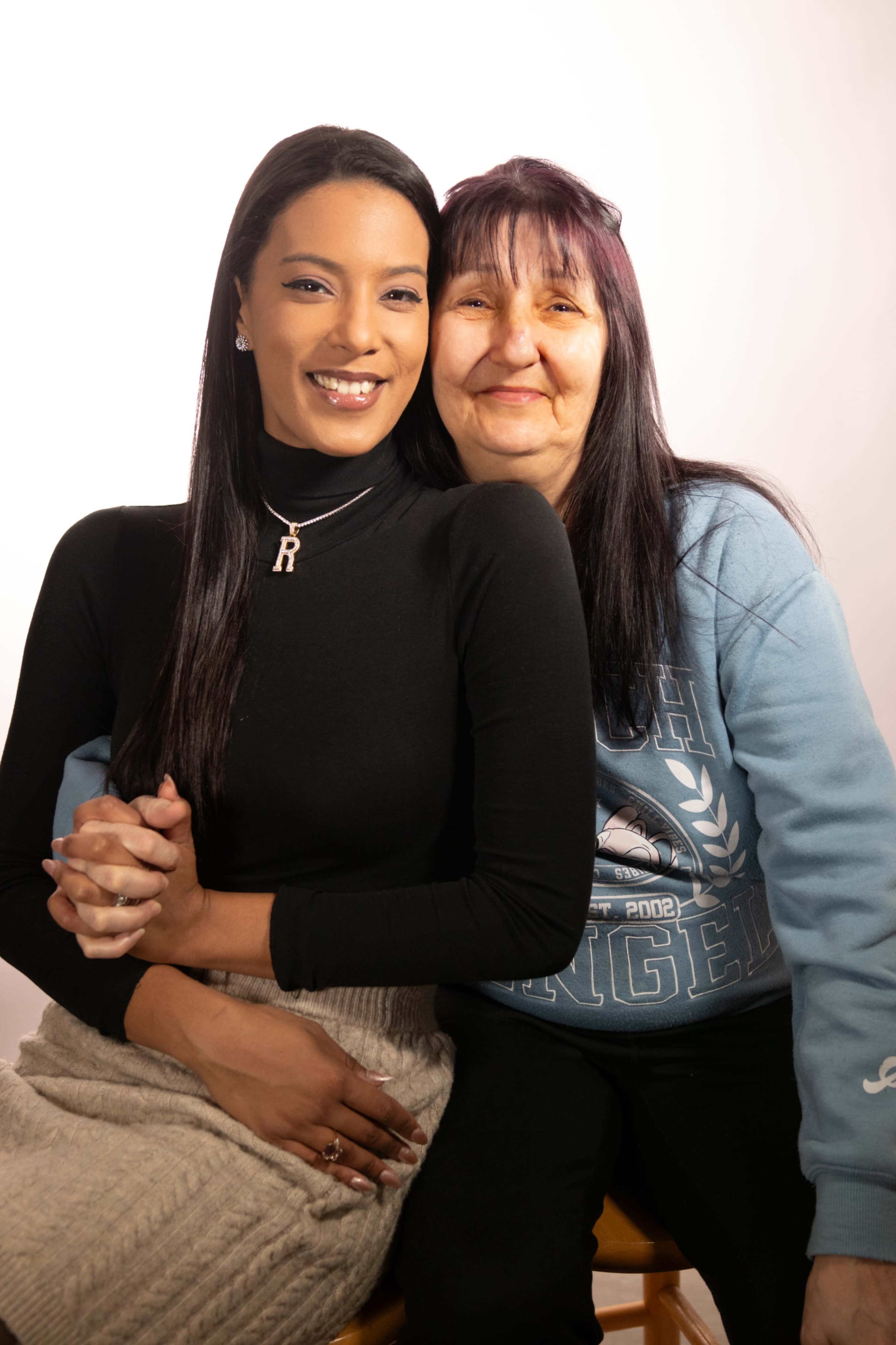 Two women sit side by side, smiling, in front of a plain background.