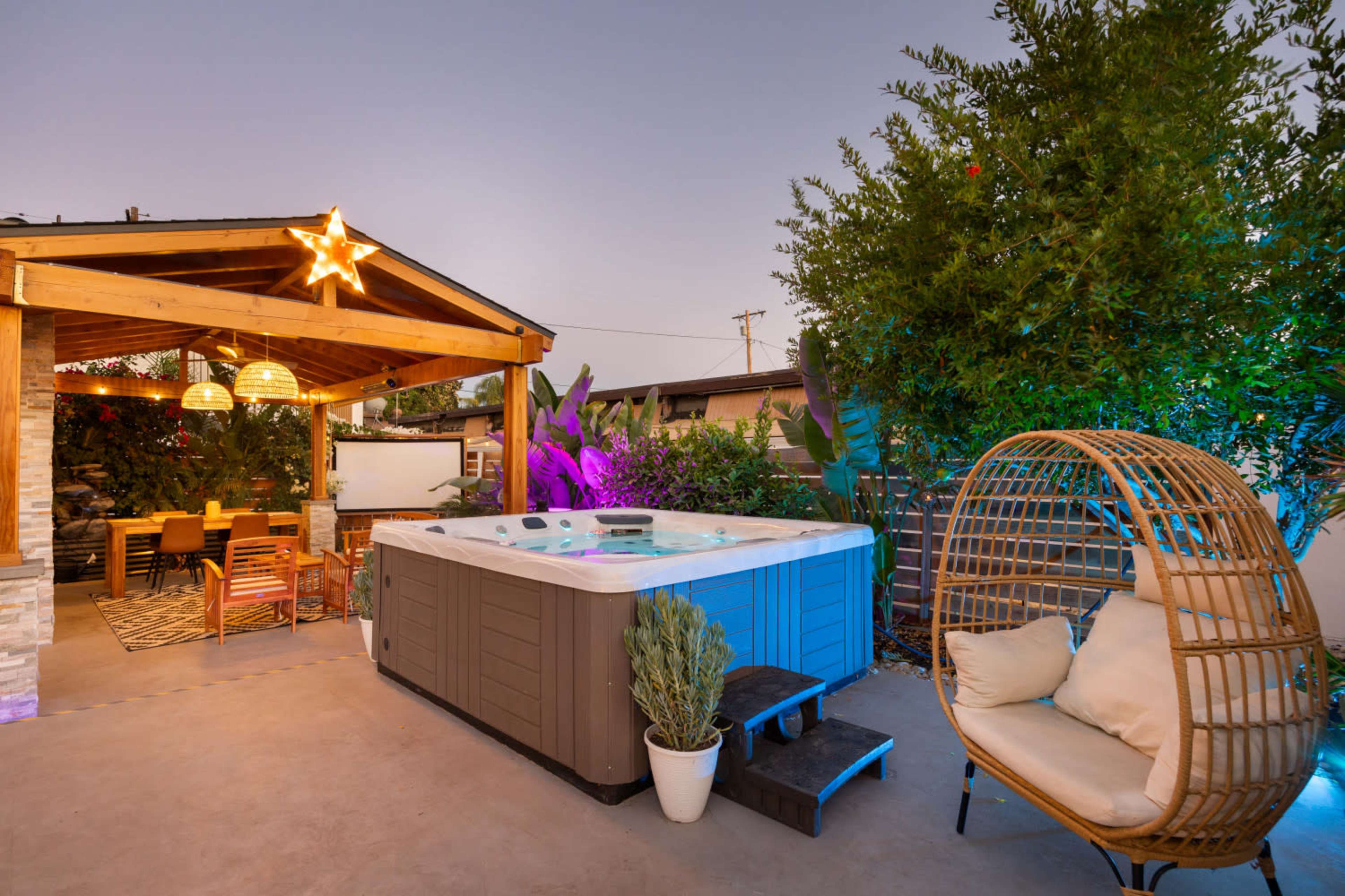 The image features a backyard scene with a hot tub, a covered seating area with a dining table, and a decorative chair surrounded by lush greenery.