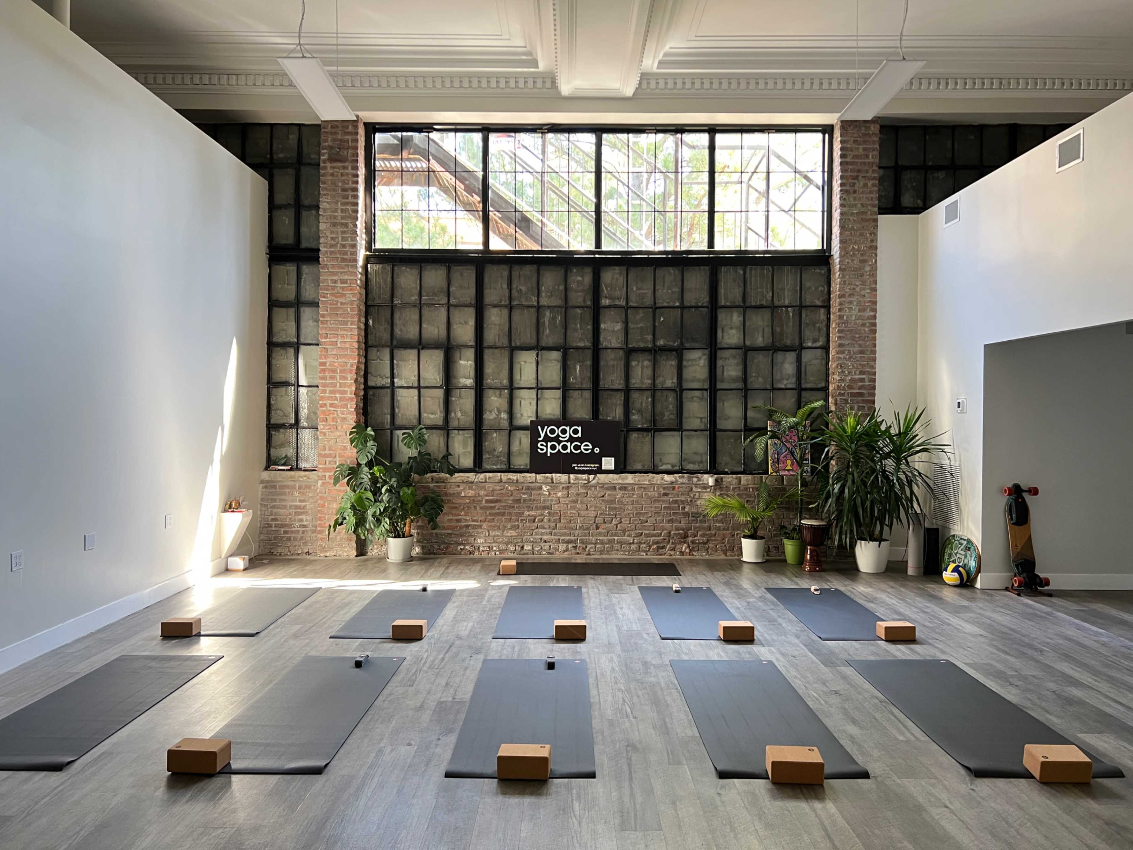 The image shows a yoga studio with several yoga mats arranged in rows, wooden blocks placed on each mat, and large windows letting in natural light.