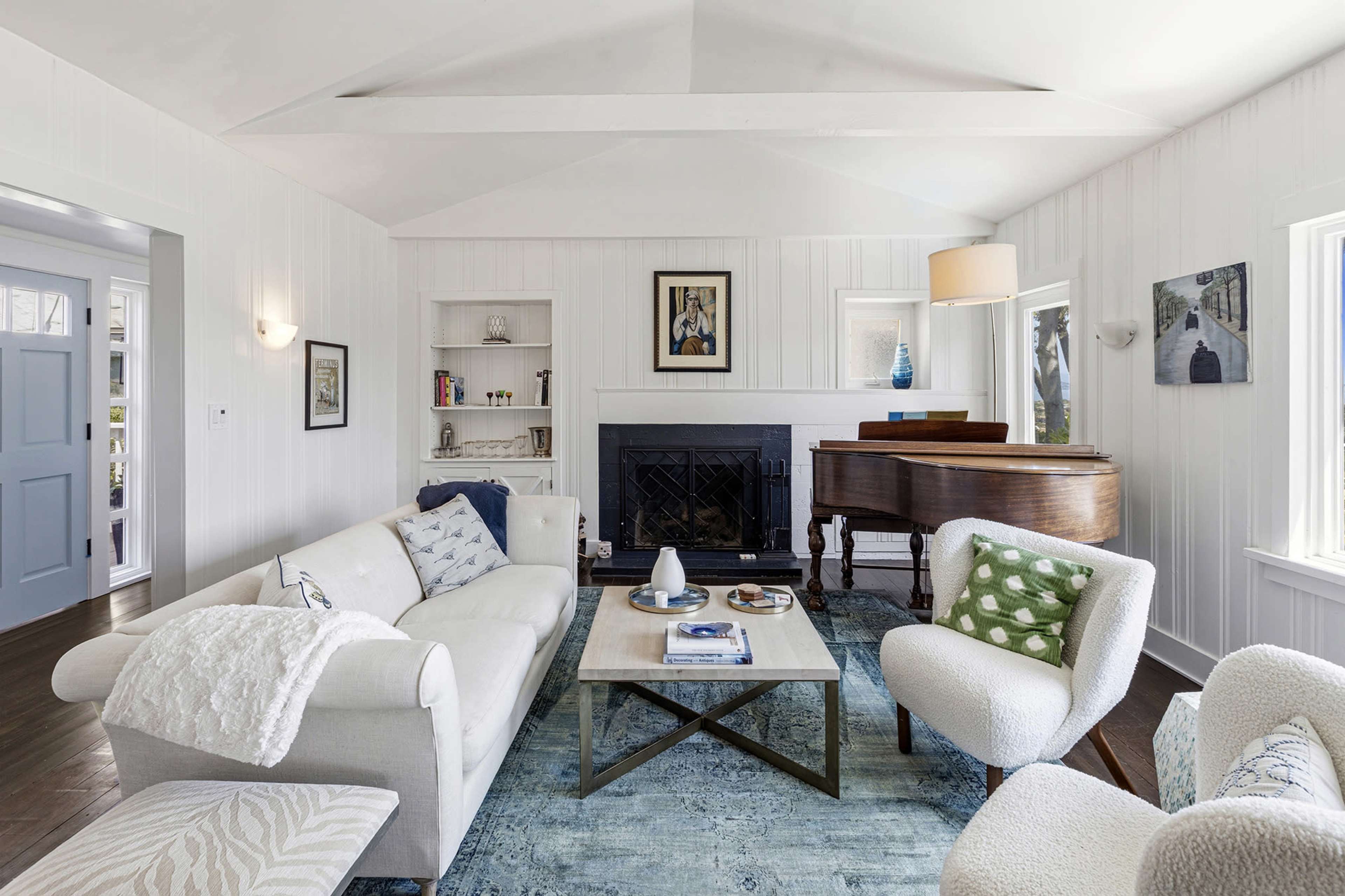 The image depicts a bright living room featuring a white sofa, two accent chairs, a coffee table, a piano, and bookshelves against light-colored walls.