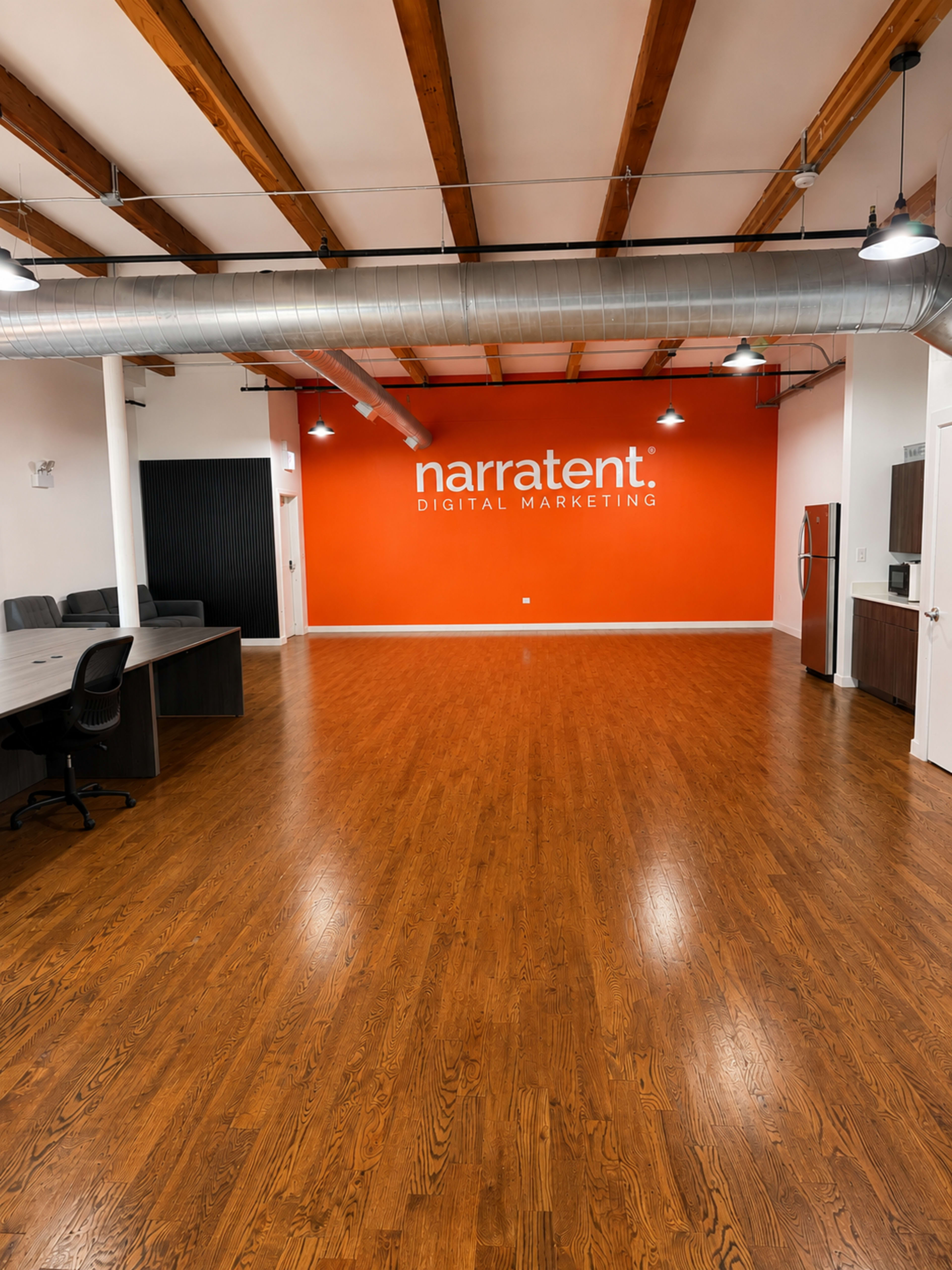 The image shows a spacious office interior with wooden flooring, exposed beams, and a bright orange wall featuring the text "narratent. DIGITAL MARKETING."