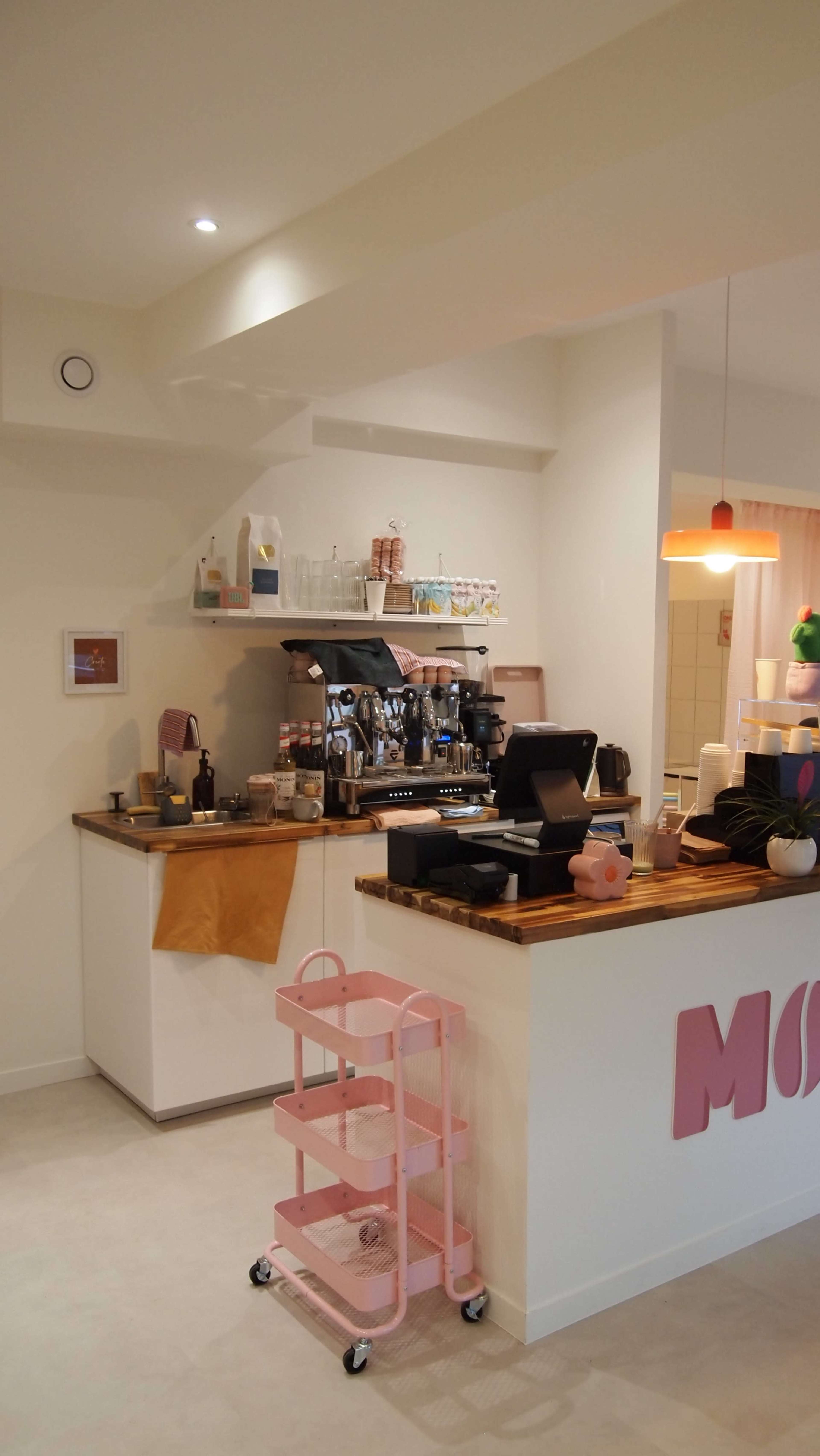 The image shows a cozy coffee shop interior with a wooden counter, a coffee machine, shelves stocked with various items, and pastel-colored decor.