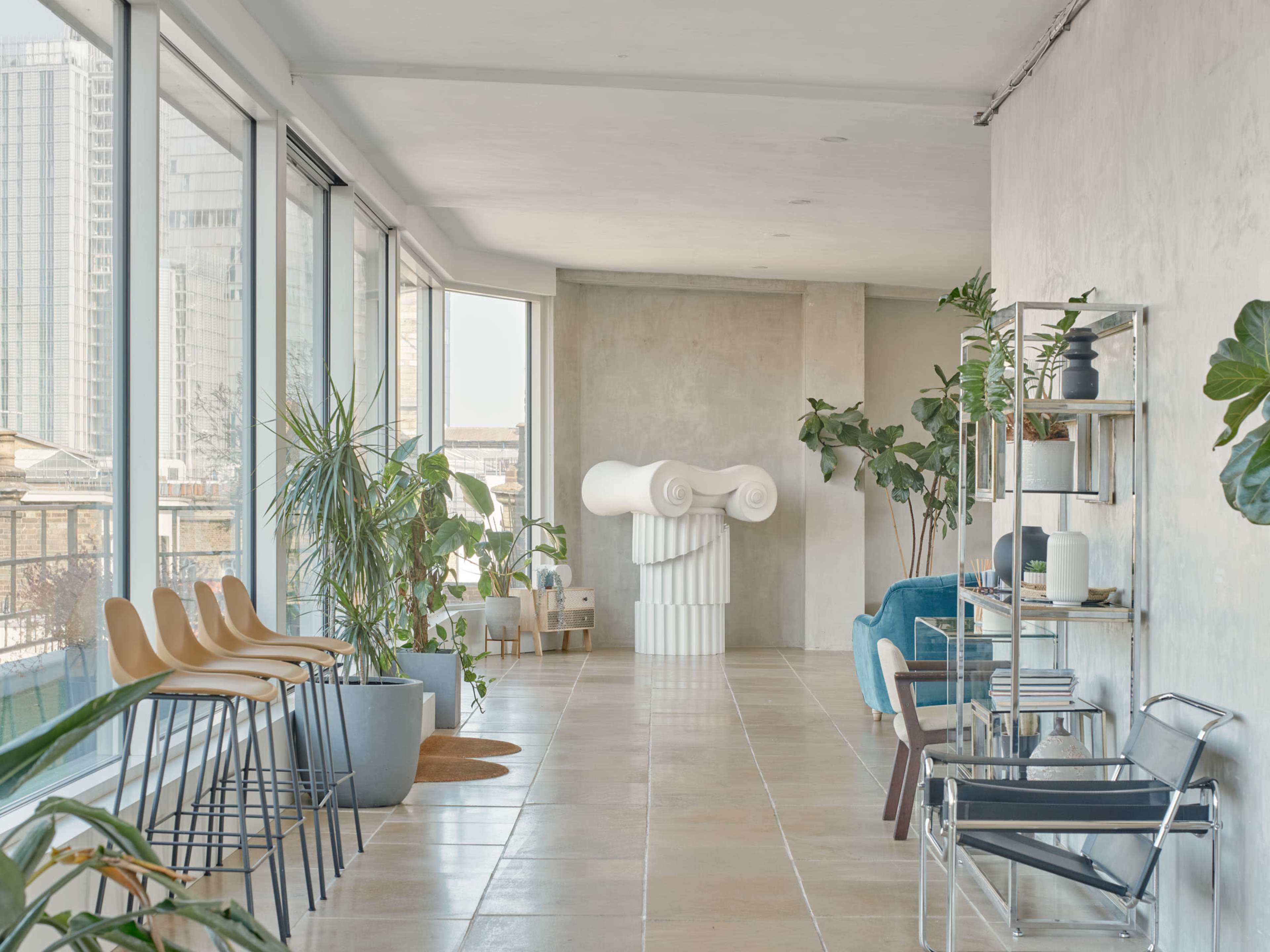 The image depicts a modern, airy interior space with large windows, featuring various plants, minimalist furniture, and an abstract sculpture at the center.