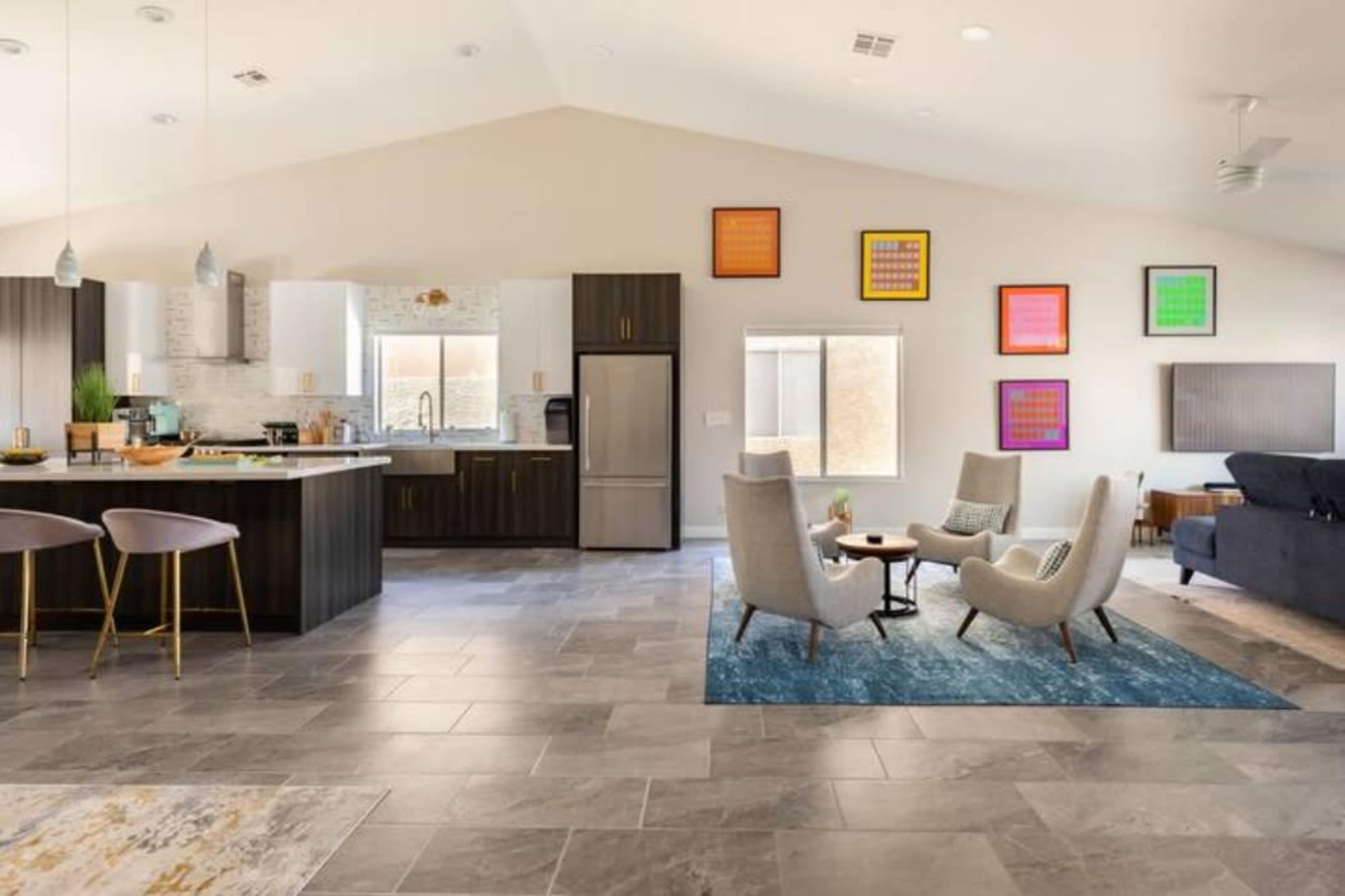 A modern open-concept living space featuring a kitchen with a dining area, a cozy seating arrangement with two armchairs, and various colorful artworks on the walls.