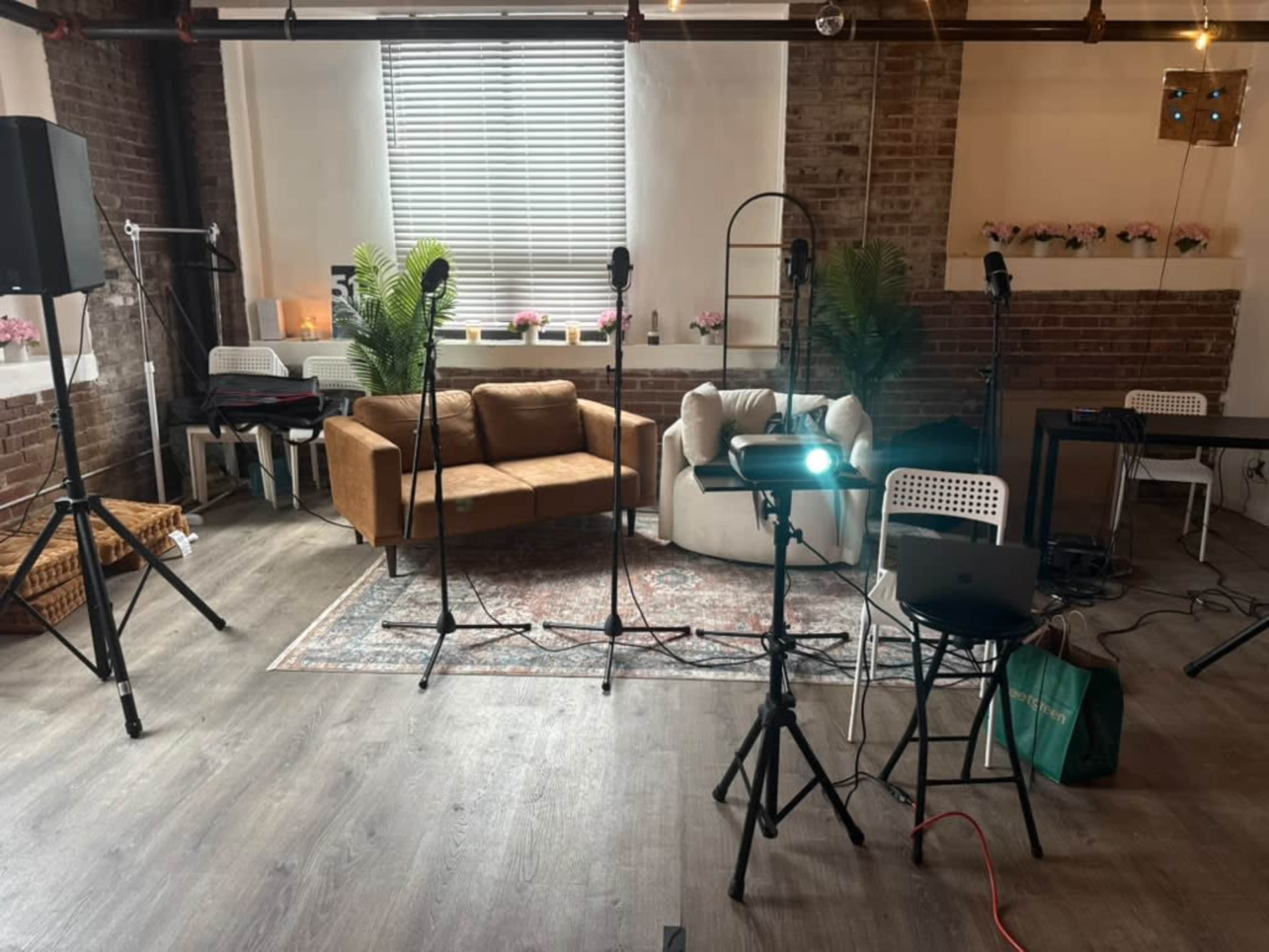 The image shows a studio space featuring a brown sofa, a small table, microphones on stands, and a projector, with wooden flooring and exposed brick walls.