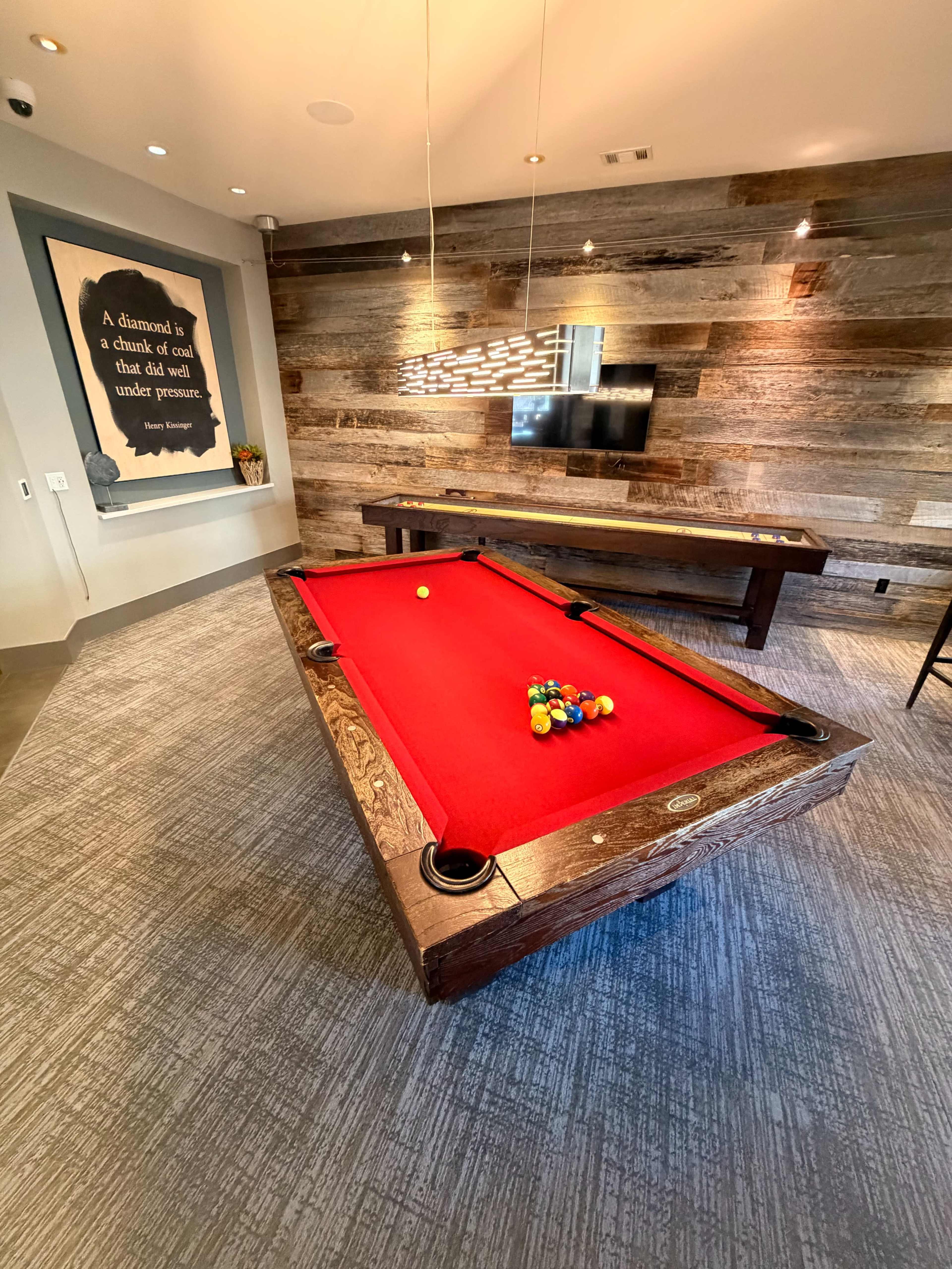The image shows a billiards room featuring a red pool table with balls racked on it, wooden walls, and a television mounted on the wall.
