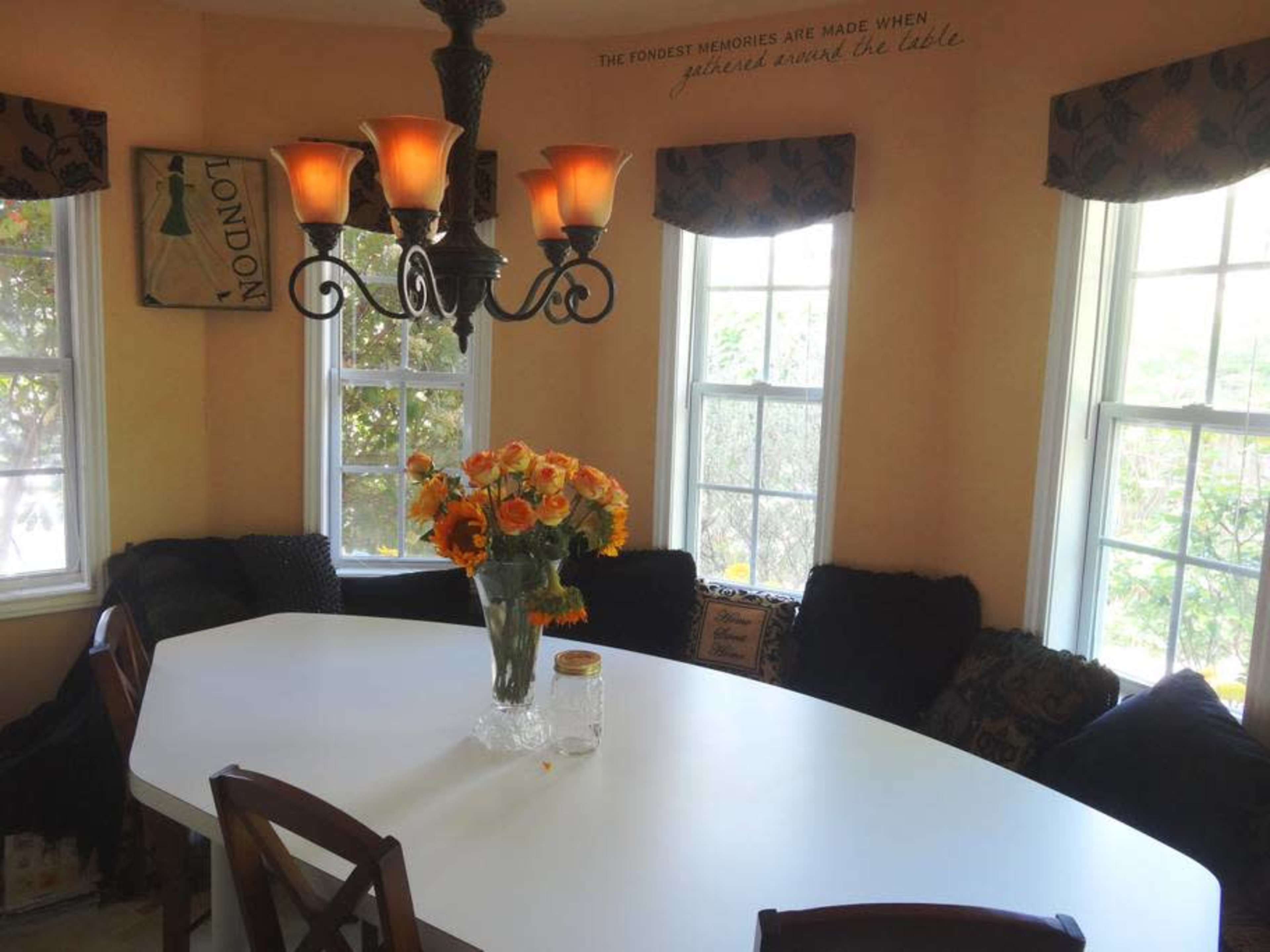 A round white dining table is surrounded by chairs in a sunny corner of a room, with large windows and a chandelier above.