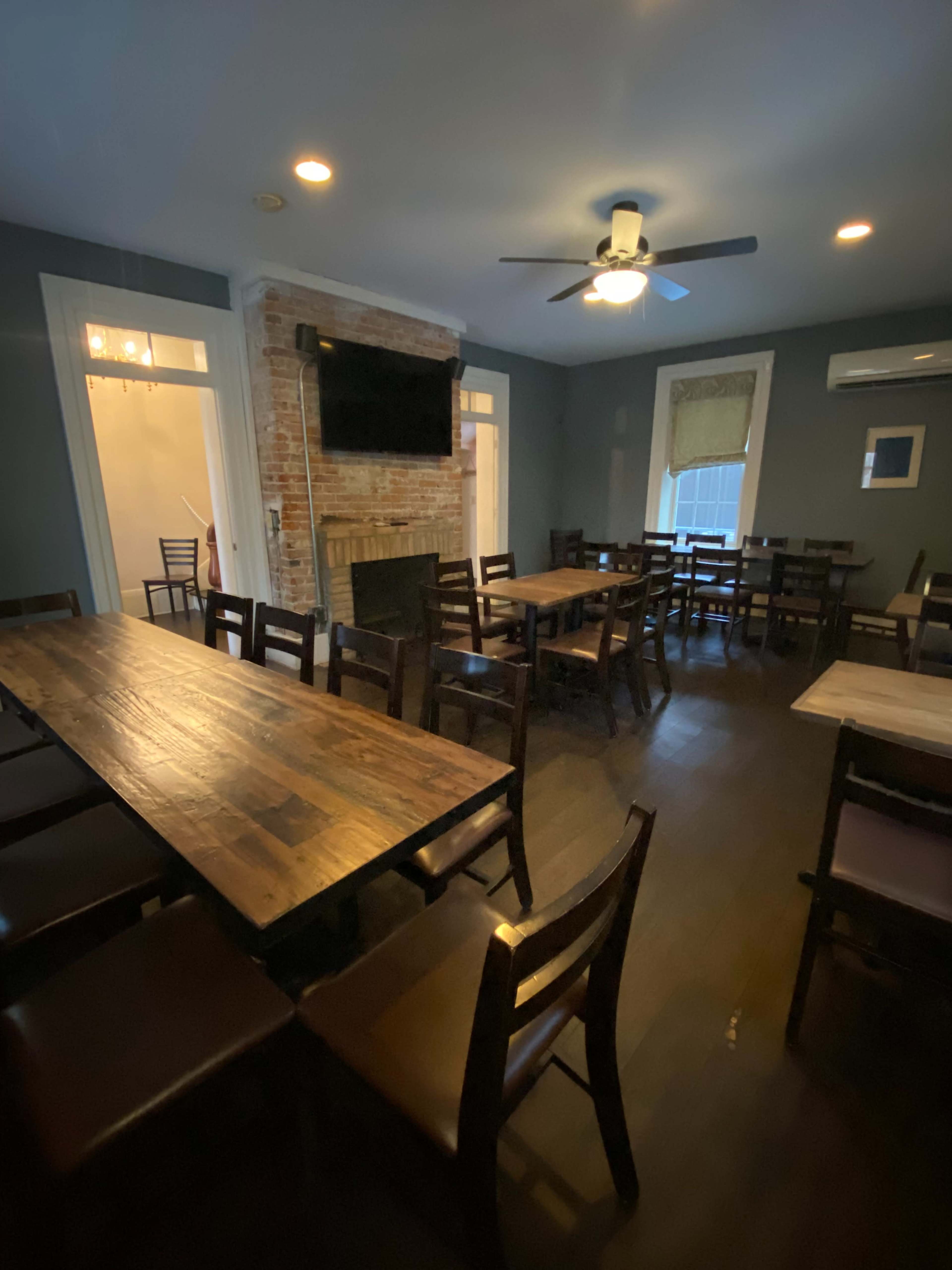 The image shows a dimly lit dining area with wooden tables and chairs arranged around a central space, featuring a brick fireplace and a ceiling fan.