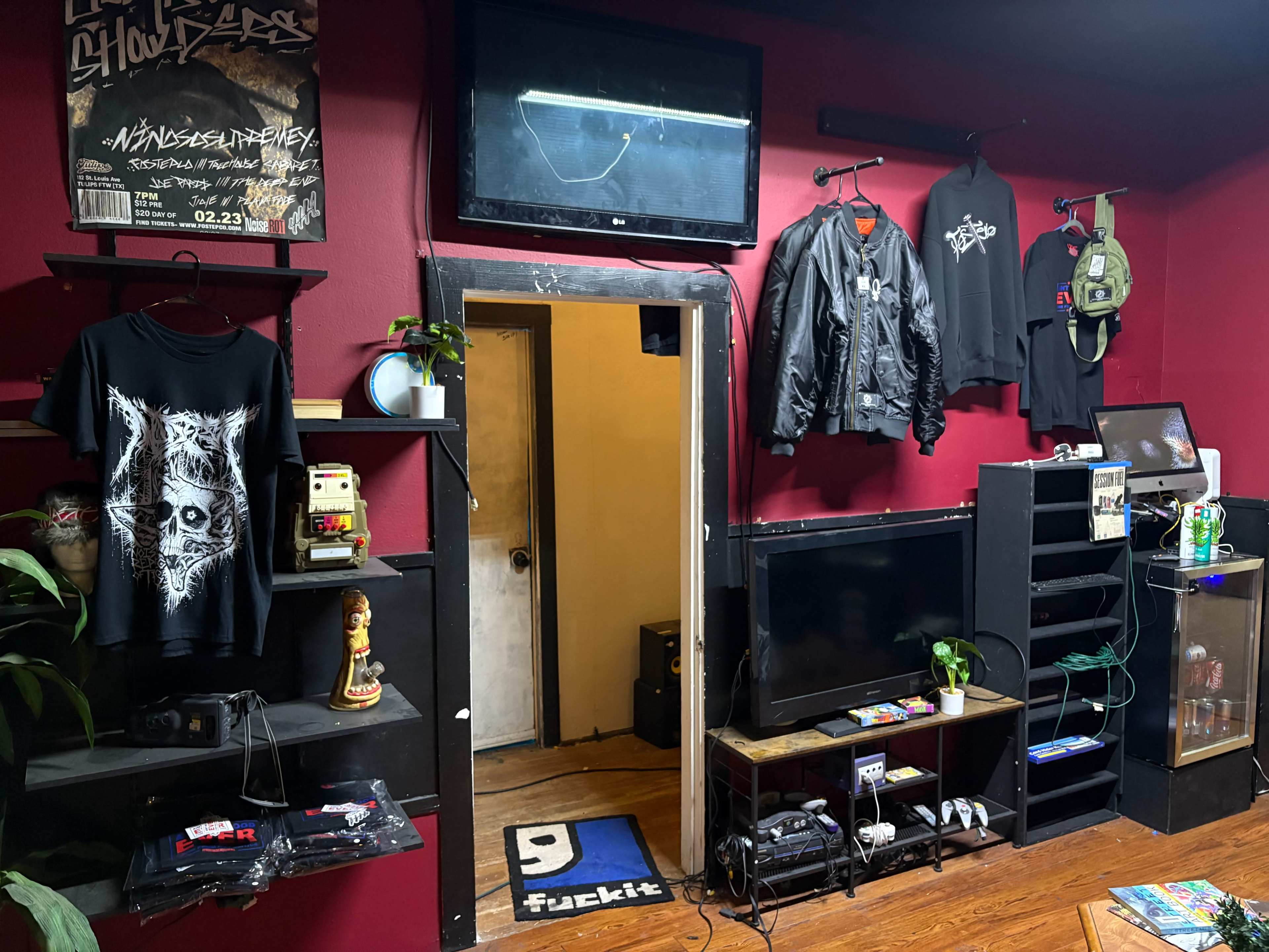 The room features a red wall adorned with clothing items and posters, a door leading to another area, and various electronics including a television and game consoles.