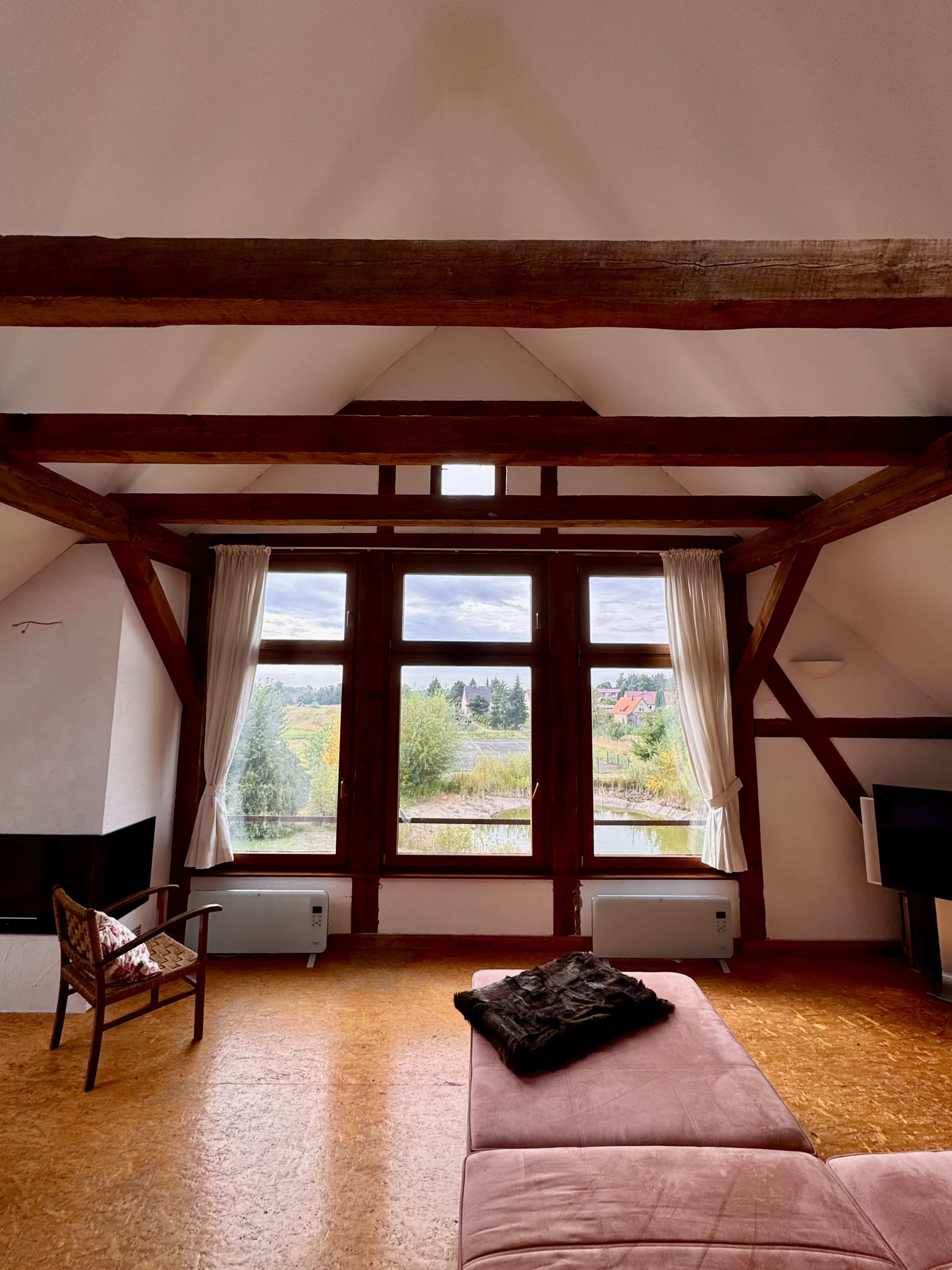 The image shows a spacious room with wooden beams, large windows overlooking a natural landscape, and minimalist furniture including a couch and a chair.